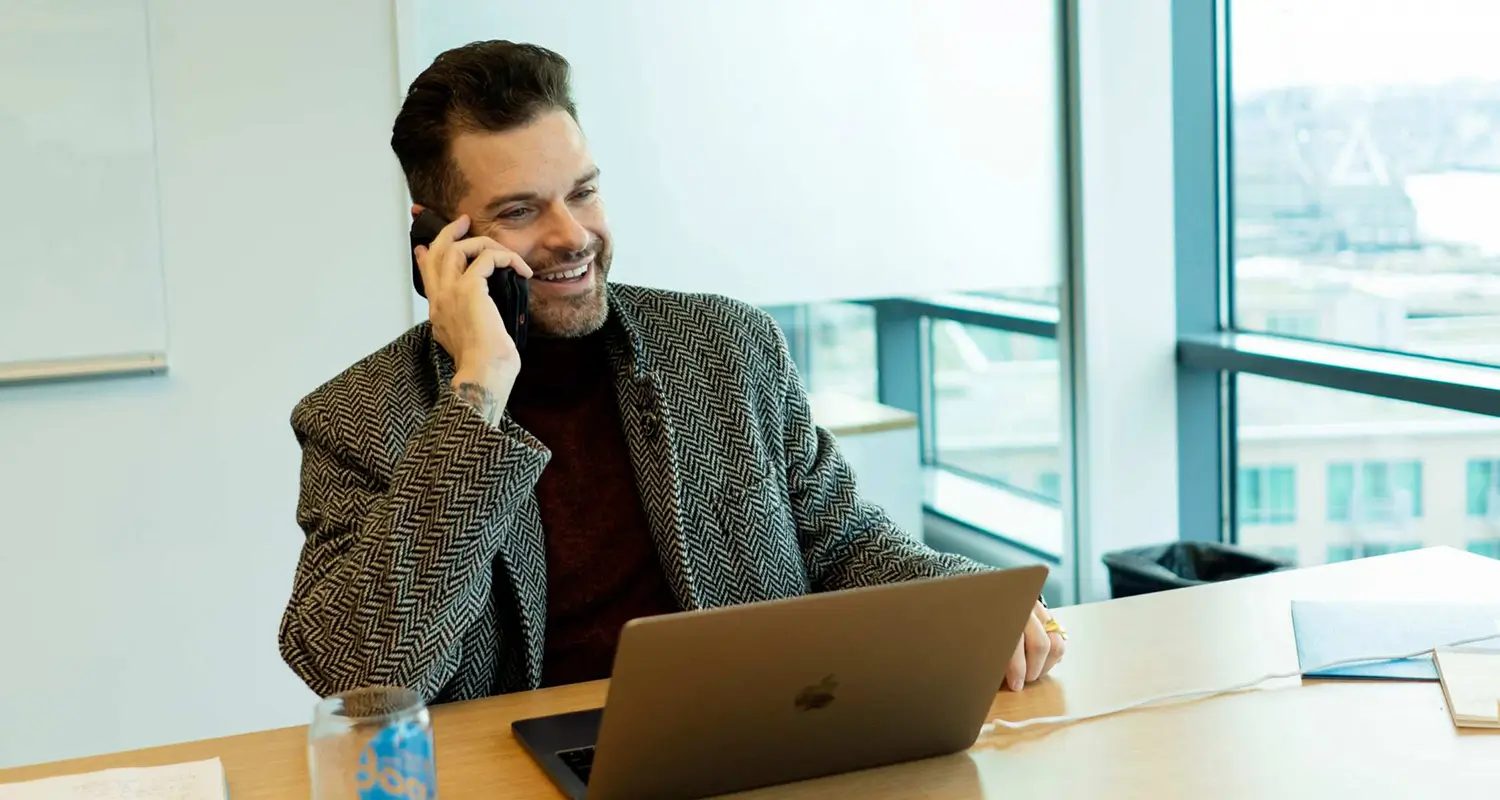 Smiling man in a patterned blazer on a phone call while working on a laptop in a modern office