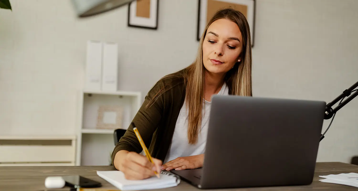 Woman with long hair writing notes in a notebook beside a laptop at a home office desk