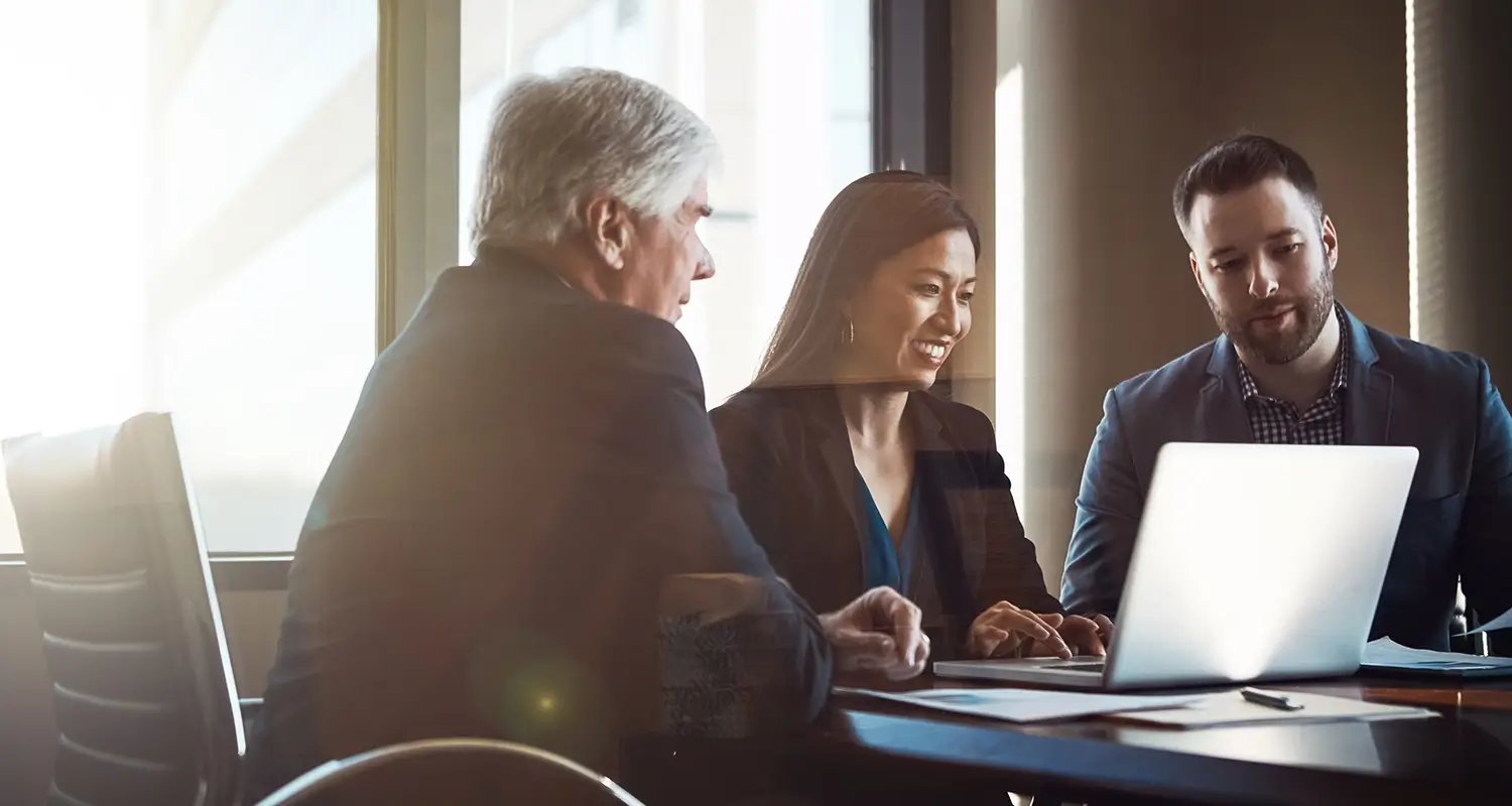 Three professionals in a meeting around a table with a laptop in a bright office setting