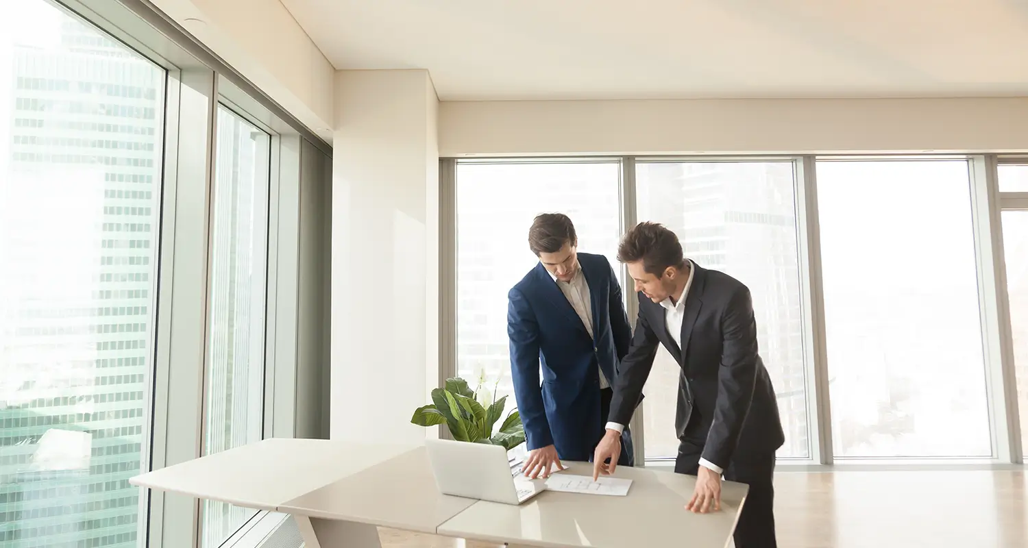 Two businessmen in suits reviewing documents on a laptop at a standing desk with city views