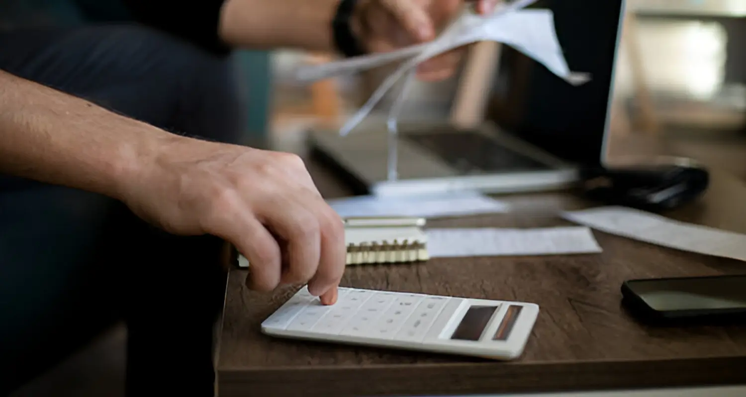 A person presses a white calculator on a wooden desk surrounded by printed financial papers, a notebook, and a smartphone, while someone else in the background sifts through additional documents. The image illustrates the detailed cost accounting process businesses undergo when uncovering the true hidden expenses embedded in their local payroll.