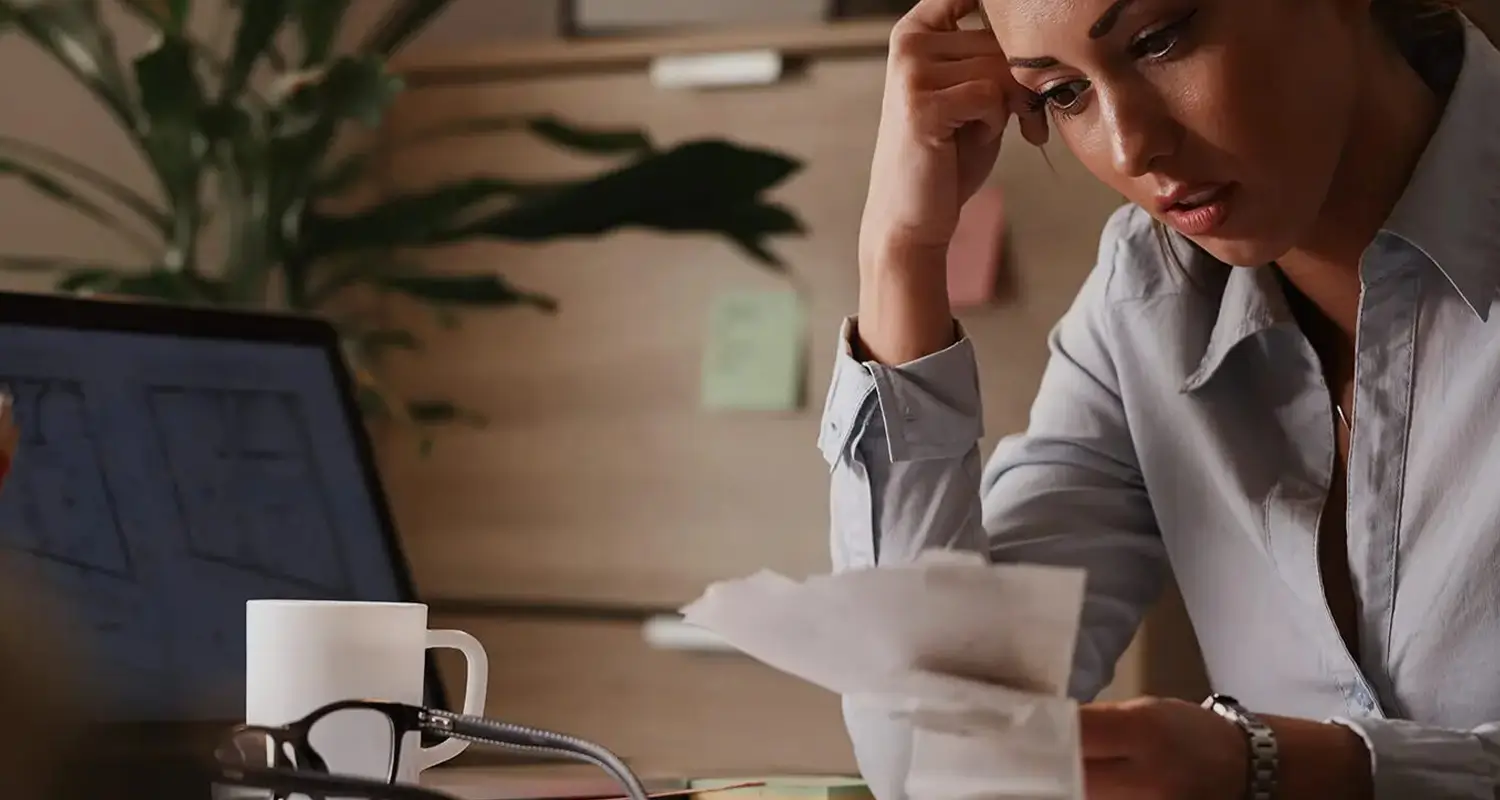 A stressed woman in a gray shirt holds her head with one hand while reviewing printed documents at a dimly lit desk, with a laptop, coffee mug, glasses, and sticky notes visible. The scene conveys the overwhelm business owners face when confronted with the full burden of hidden payroll costs beyond base salaries.