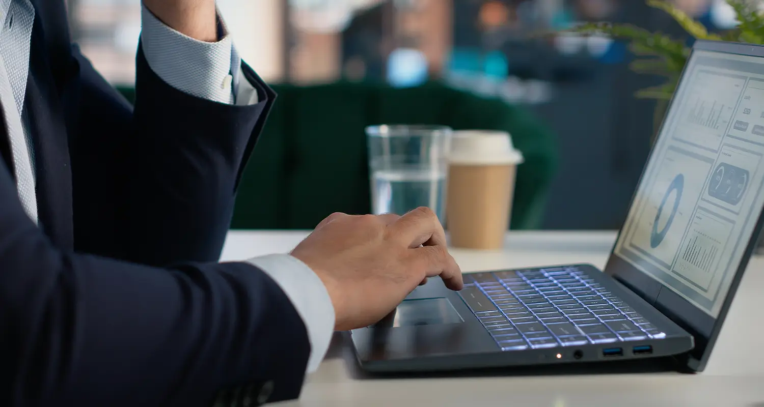 A close-up of a business professional's hands typing on a glowing laptop keyboard, with a dashboard showing pie charts and bar graphs visible on the screen. A takeaway coffee cup and water glass sit nearby, representing the data-driven operational oversight a MaaS leader exercises to monitor and manage a globally distributed team that saved over $900K.