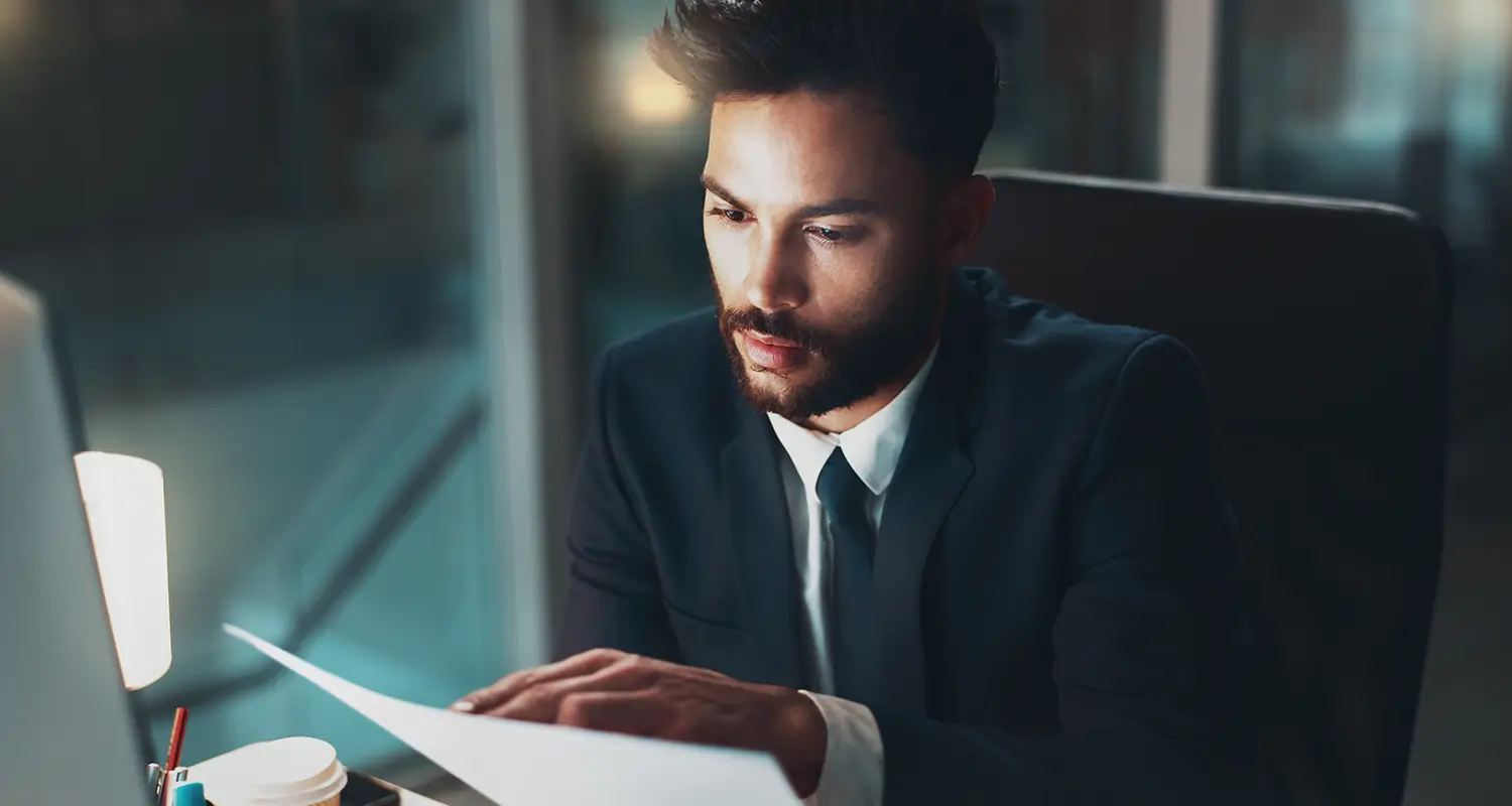A professional man in a dark suit reads a printed document intently at a desk illuminated by a side lamp in a dimly lit office with glass partitions visible in the background. The focused, late-night setting represents a calibration industry leader reviewing business reports as part of the operational decision-making that led to building an offshore team and saving over $630K.