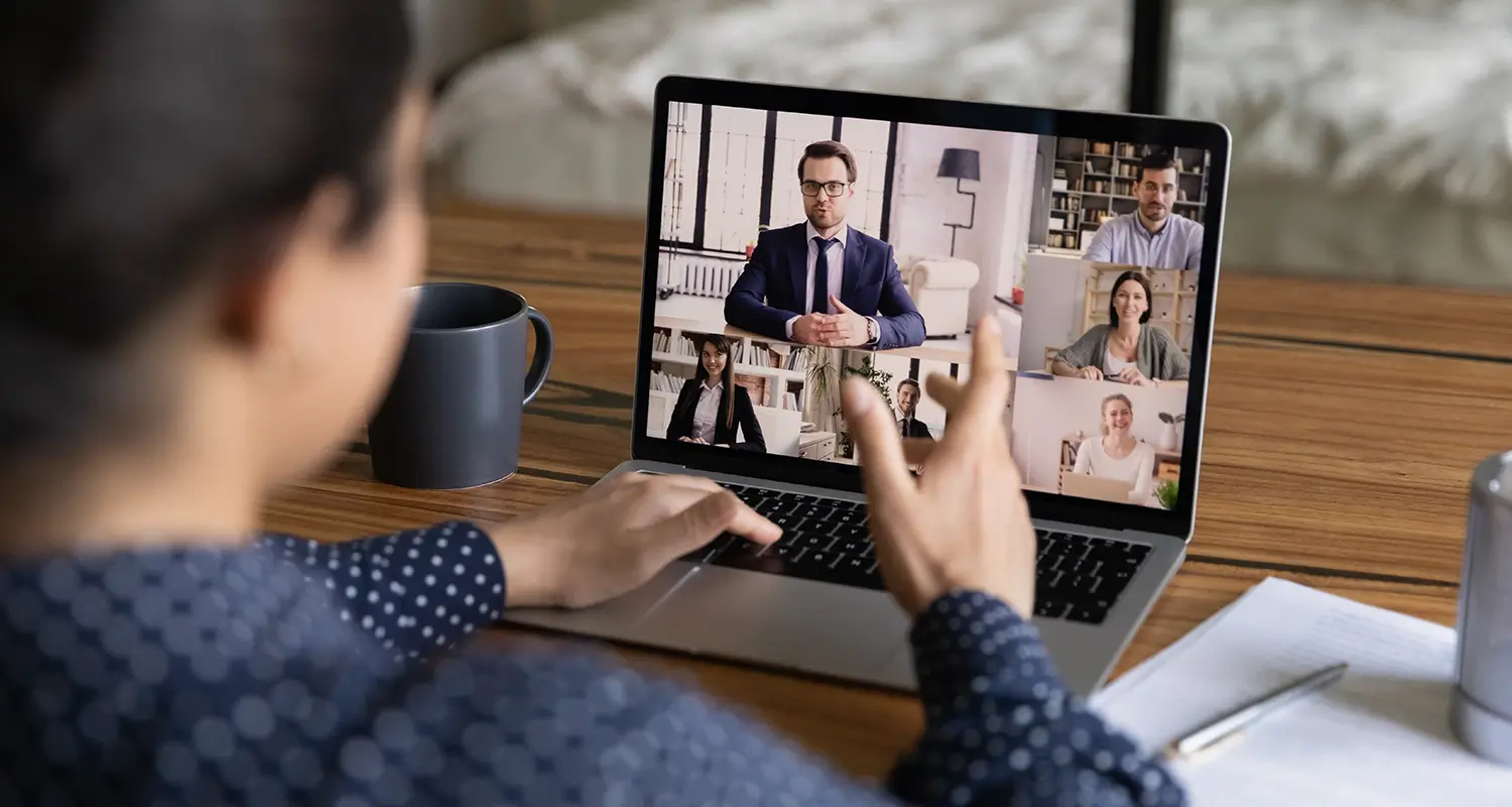 A woman seen from behind gestures toward a laptop screen showing a five-person video conference call with professionals in various home and office settings, with a coffee mug and notebook on the desk. The image illustrates the remote interview and vetting processes central to offshore recruitment services, helping companies build distributed teams efficiently.