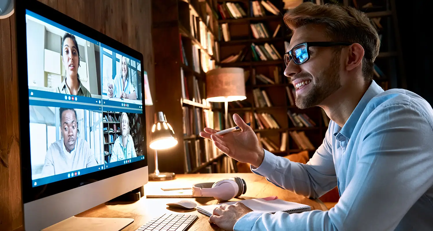 A smiling young man with glasses sits at a large desktop monitor showing a four-person video call grid, with a warm bookshelf background visible behind him as he gestures expressively with a pen in hand. The scene represents a recruiter or hiring manager using offshore recruitment services to engage and evaluate global candidates through structured virtual interviews.