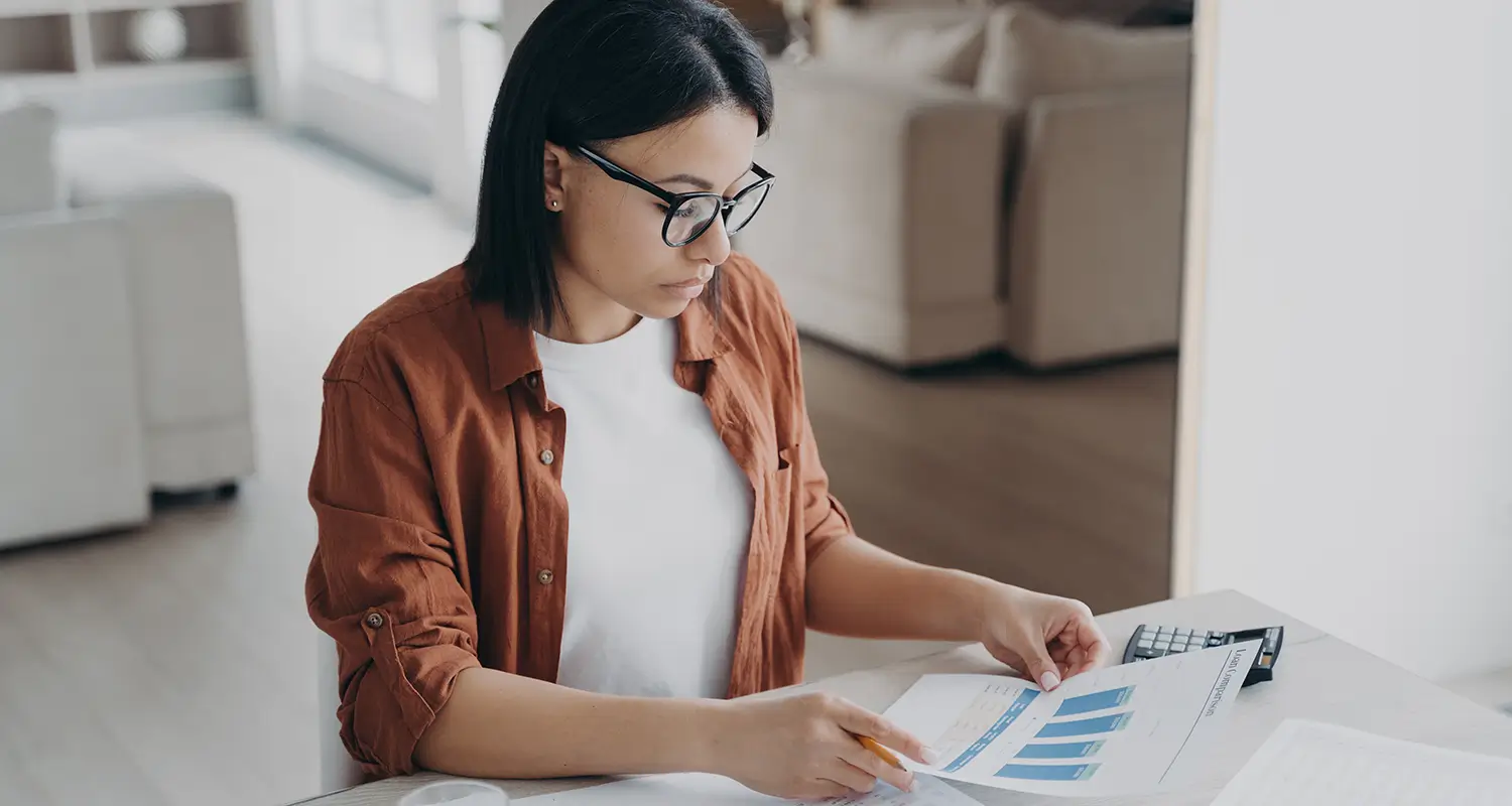 A young woman with dark hair and glasses sits at a table, attentively reviewing a printed report with bar charts while operating a calculator. The focused, analytical posture represents an offshore construction estimator or cost analyst carefully reviewing project financials to help construction companies improve bid accuracy and profitability.