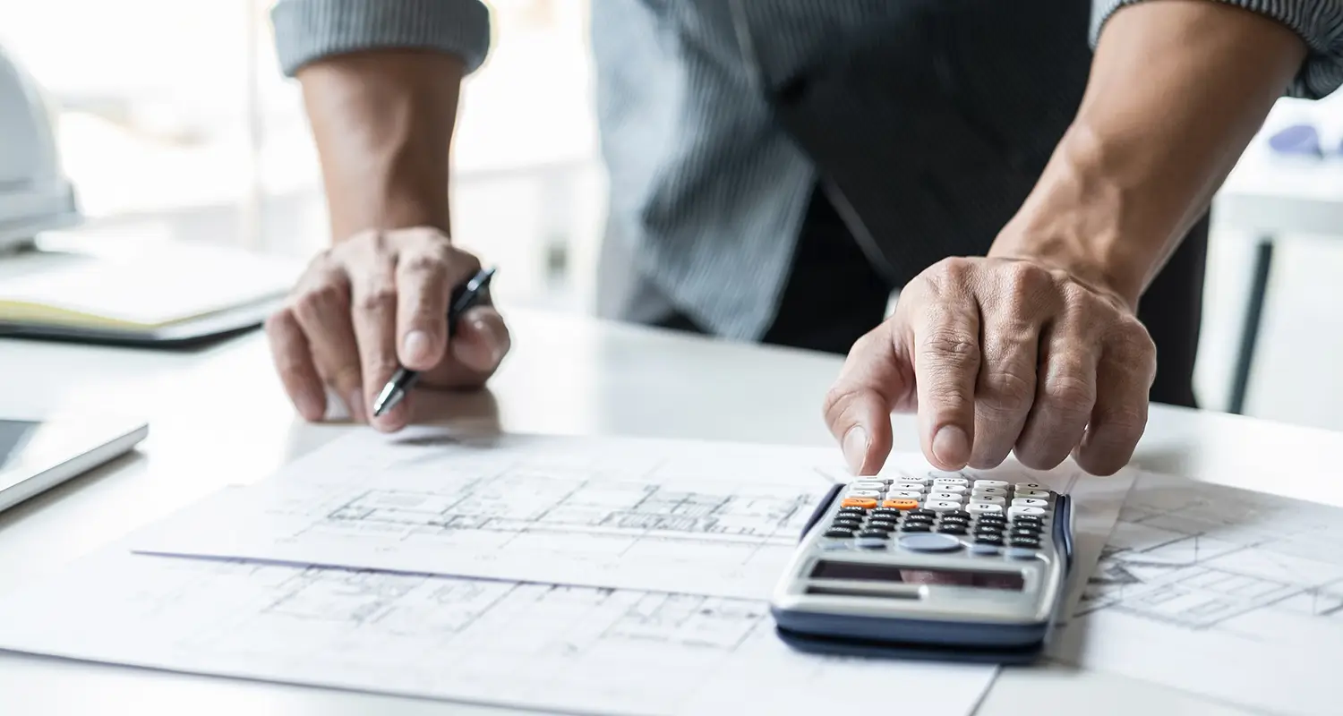 A close-up of two hands working at a desk covered in architectural blueprints and construction plans, with one hand operating a calculator and the other holding a pen. The image directly represents the core work of an offshore construction estimator: performing precise quantity takeoffs and cost calculations from project drawings to support competitive bid submissions.