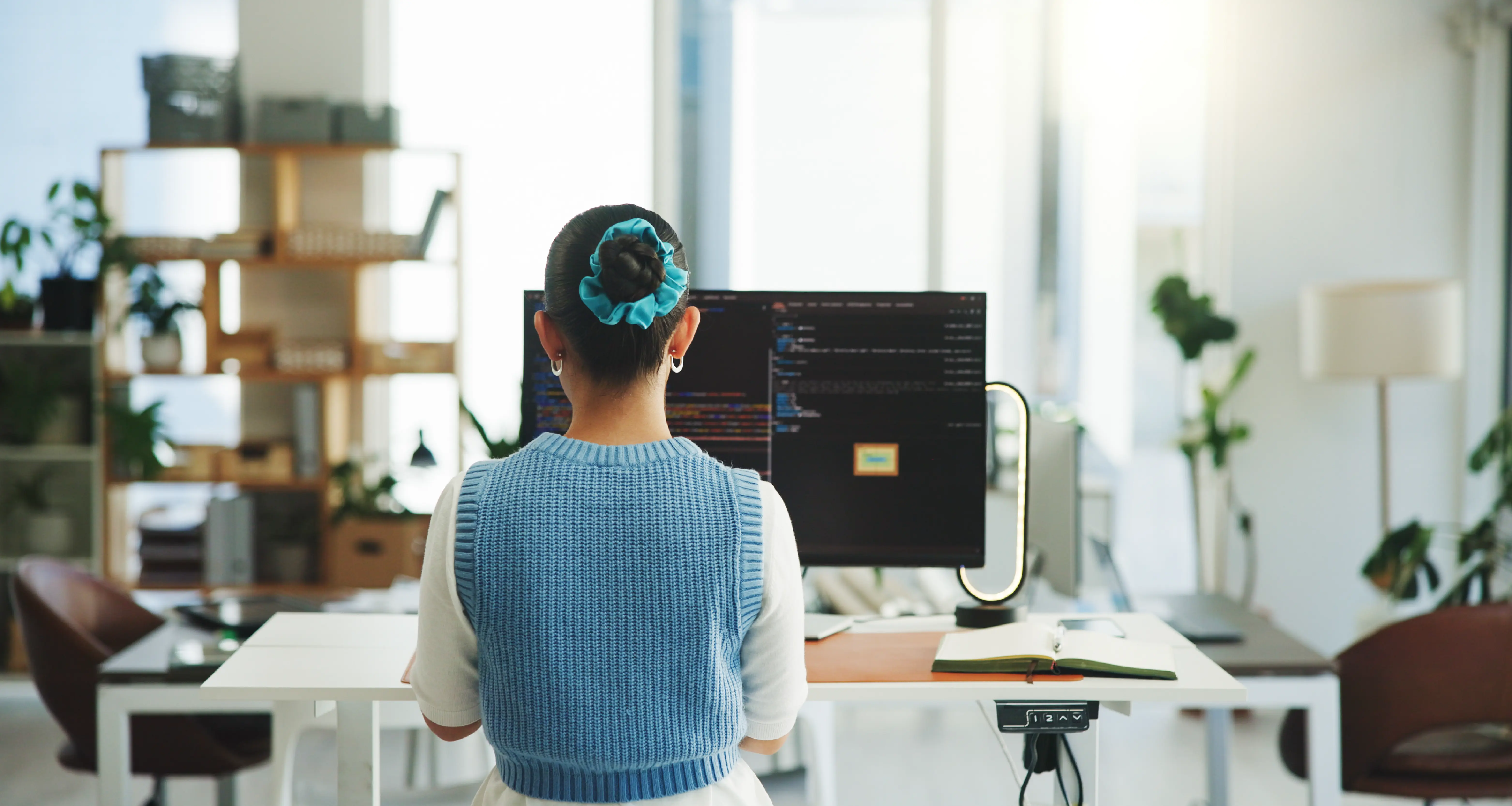 A software engineer with a blue scrunchie sits at a standing desk with her back to the camera, facing a large monitor displaying lines of code in a dark-themed IDE. The open-plan workspace behind her features shelving, houseplants, and natural light.