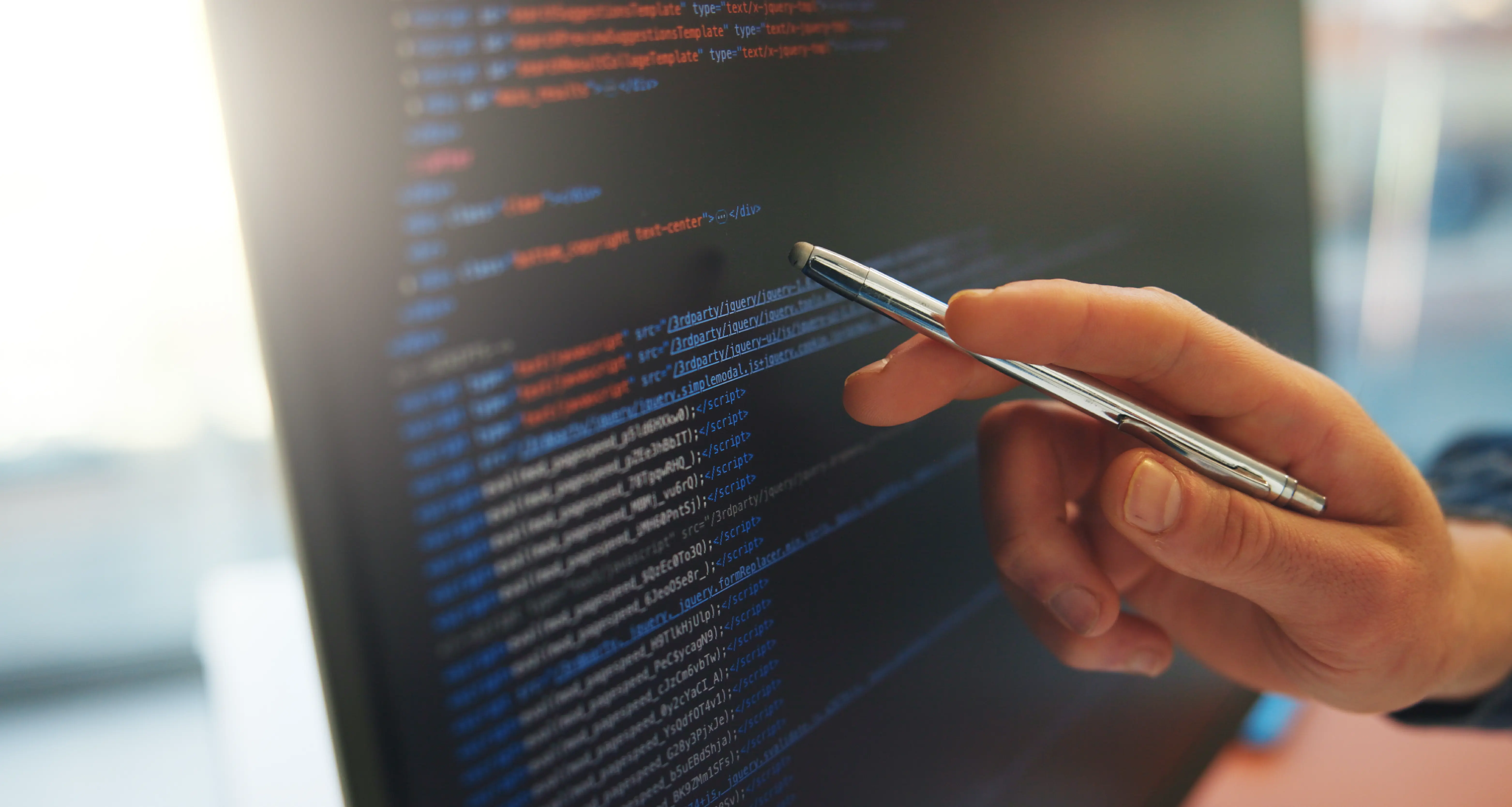 A close-up of a hand holding a pen pointing at a dark monitor screen filled with HTML and JavaScript code displayed in blue, orange, and white syntax highlighting, suggesting a developer reviewing or debugging code.