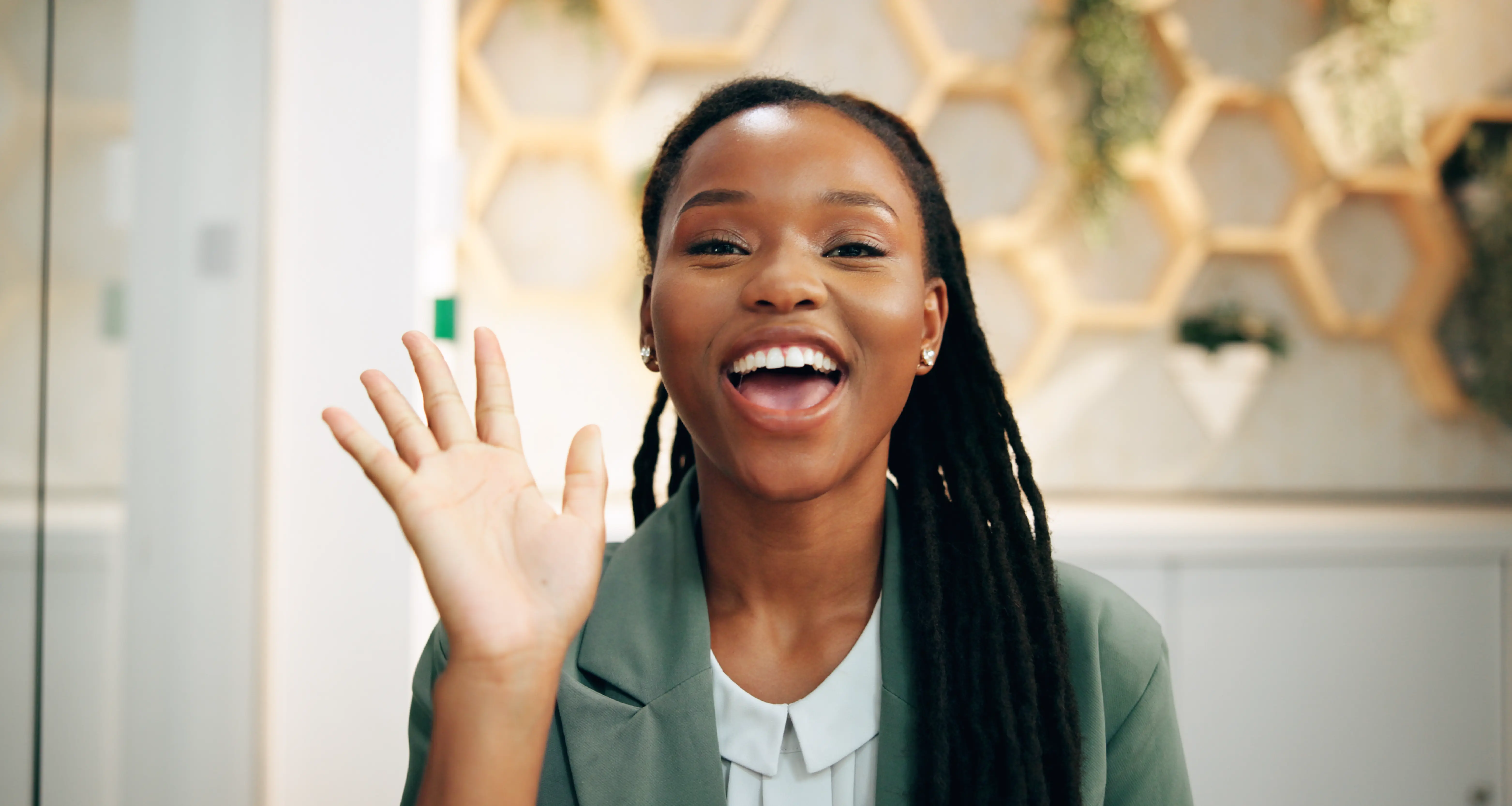 A young Black woman in a sage green blazer and braided hair waves enthusiastically and smiles broadly at the camera in a stylish, light-filled hospitality space with honeycomb wall decor. She represents an offshore team member delivering warm, high-quality guest service and communication for a luxury hospitality brand that saved over $480K through global hiring.