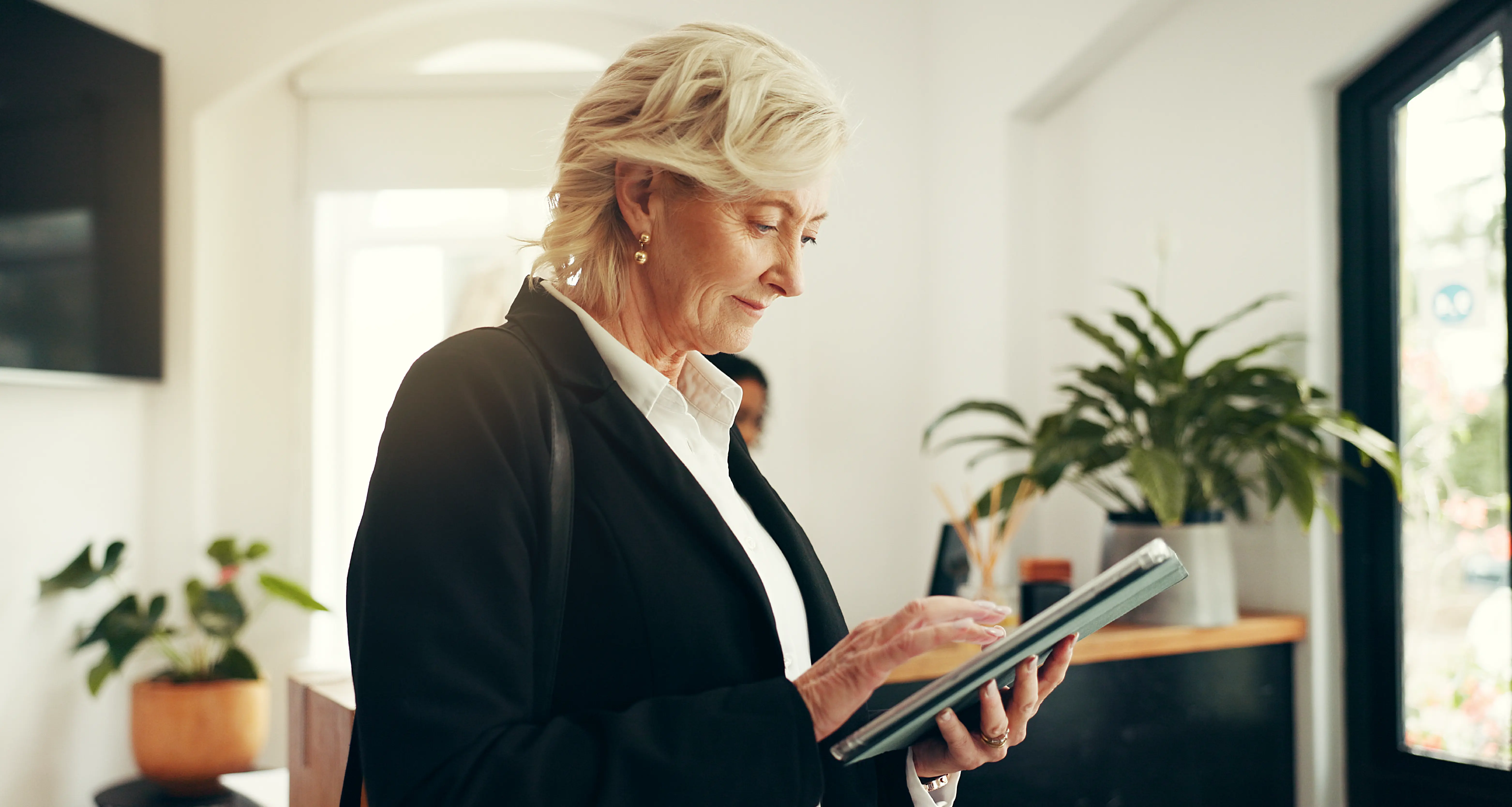 An elegantly dressed senior woman with short silver hair stands in the bright, plant-filled lobby of what appears to be an upscale hotel or hospitality venue, scrolling through content on a tablet. Her polished appearance and calm authority represent the leadership style of a luxury hospitality executive managing an offshore operations team.