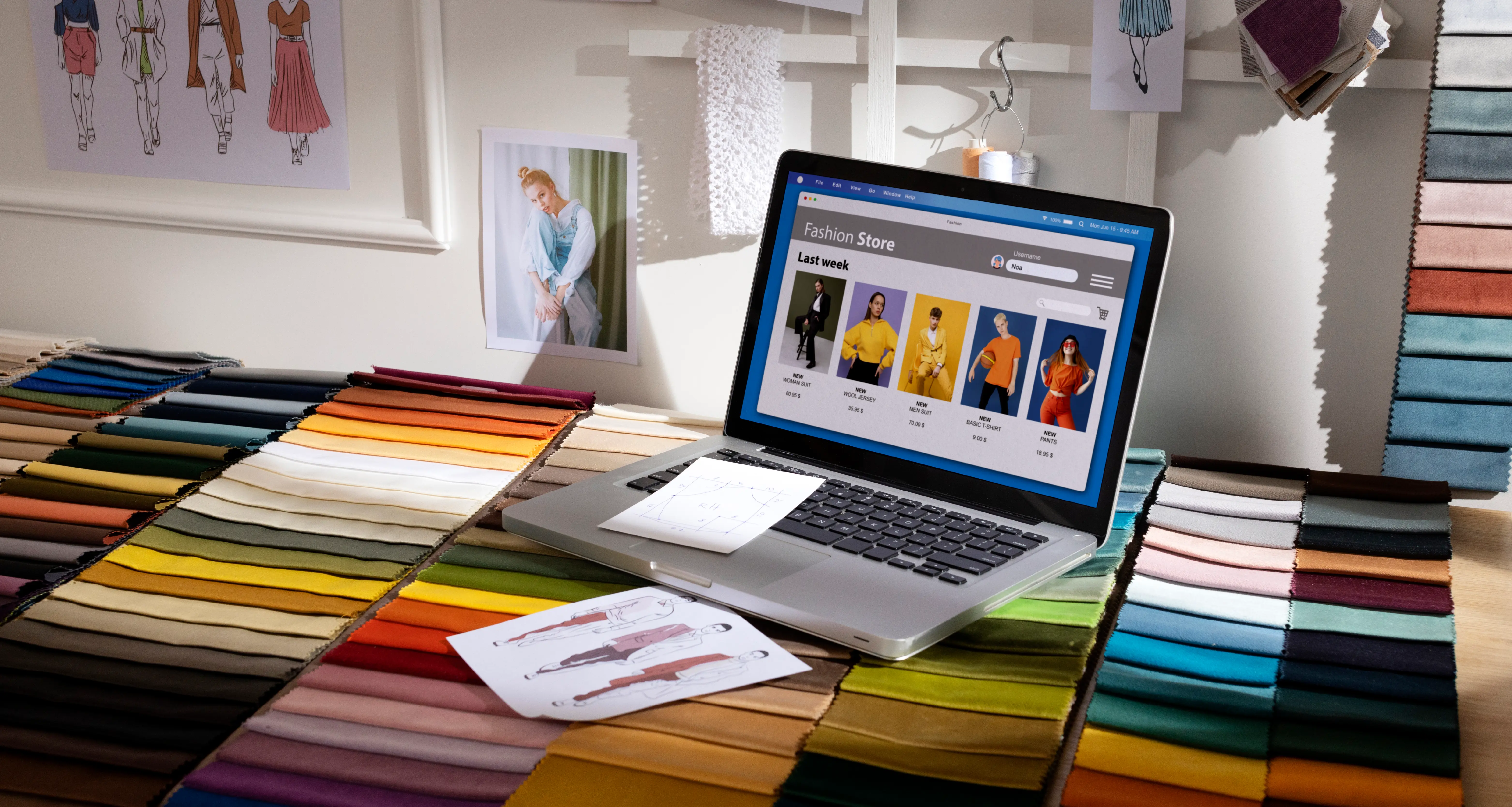 A laptop displaying a "Fashion Store" e-commerce website with a "Last week" section showing product listings sits atop a colorful spread of fabric swatches in a fashion design studio. Clothing sketches and mood board photos are pinned to the wall in the background.