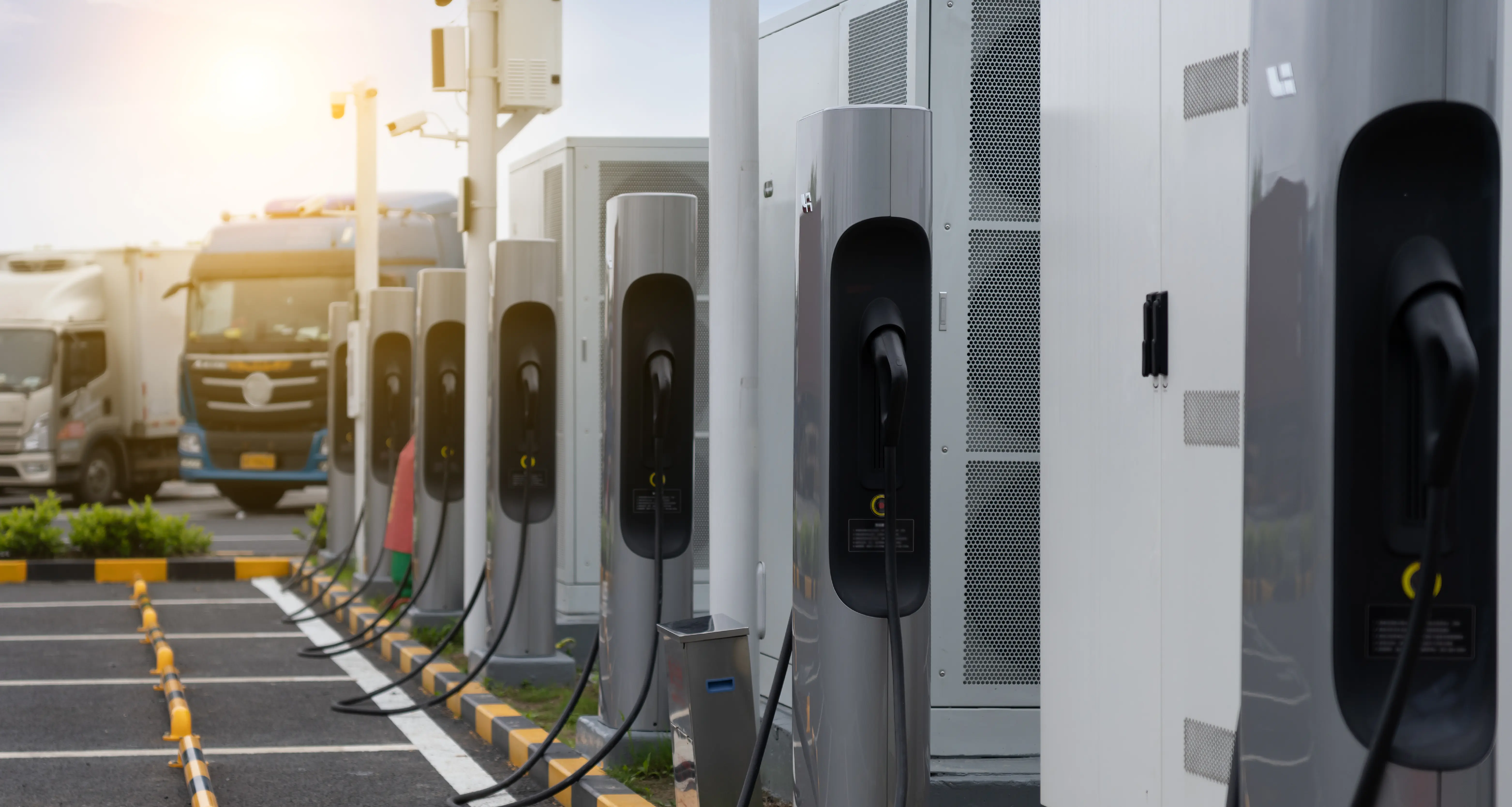 A row of commercial electric vehicle charging stations lines a paved lot at sunrise, with cables plugged in and large trucks parked in the background. The infrastructure illustrates a fleet electrification depot or charging hub.