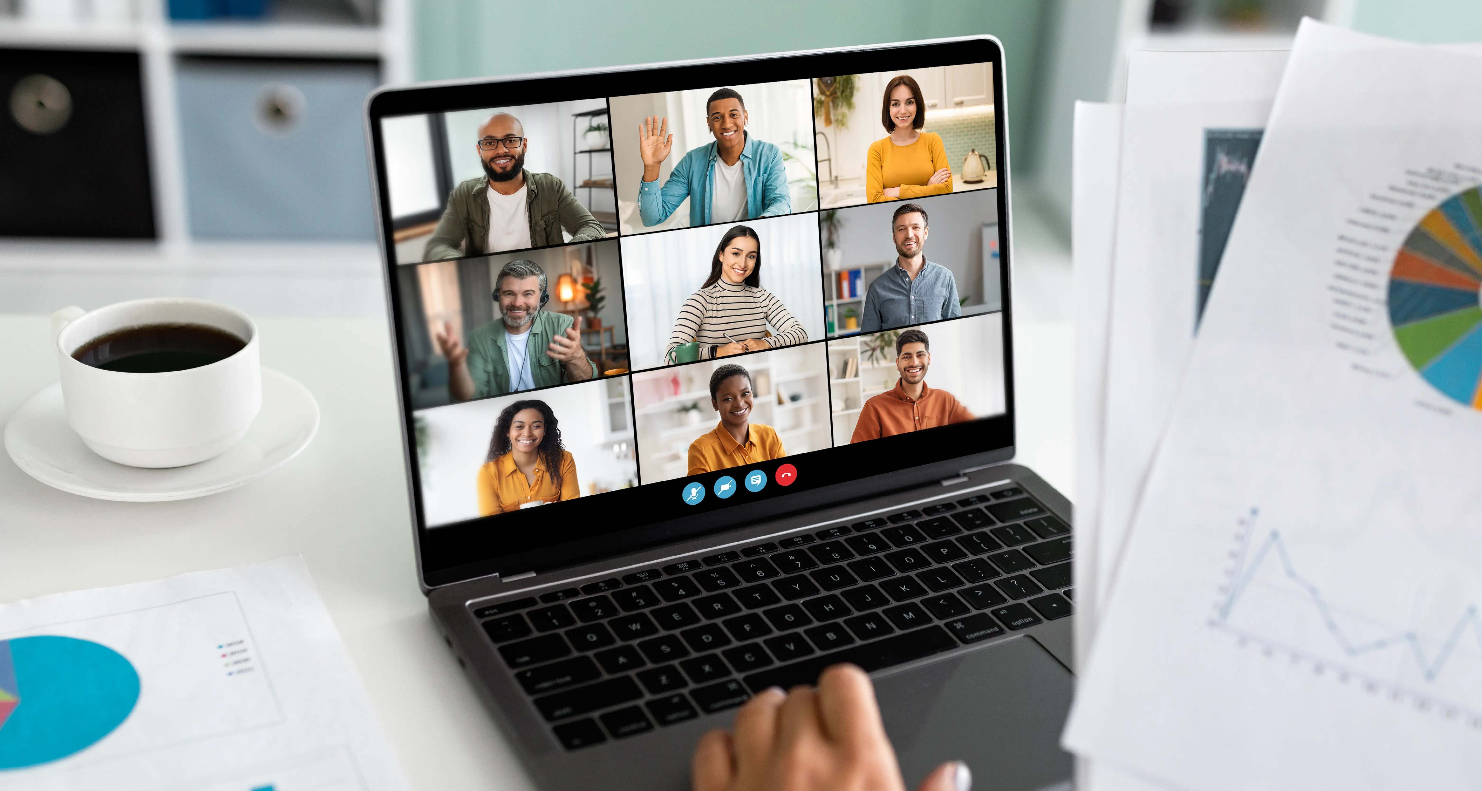 A laptop screen displays a video call with nine diverse team members in a grid layout, some waving and smiling. Printed charts and a coffee cup are visible on the desk beside the laptop, reflecting a distributed remote team meeting.