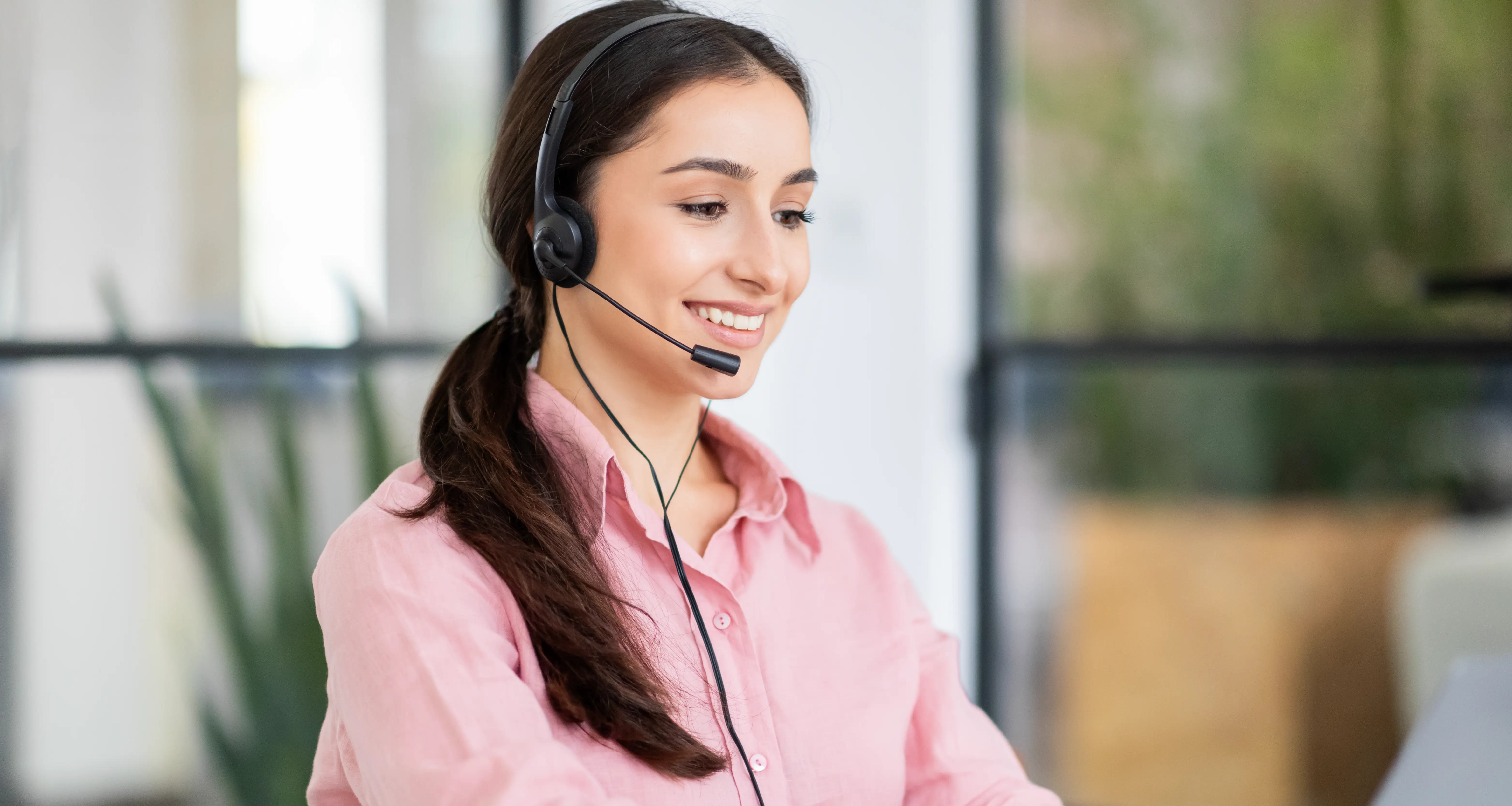 A smiling young woman wearing a headset with a microphone looks down at a screen while working in a bright, plant-filled office. Her pink shirt and professional demeanor suggest a remote assistant or customer support role.