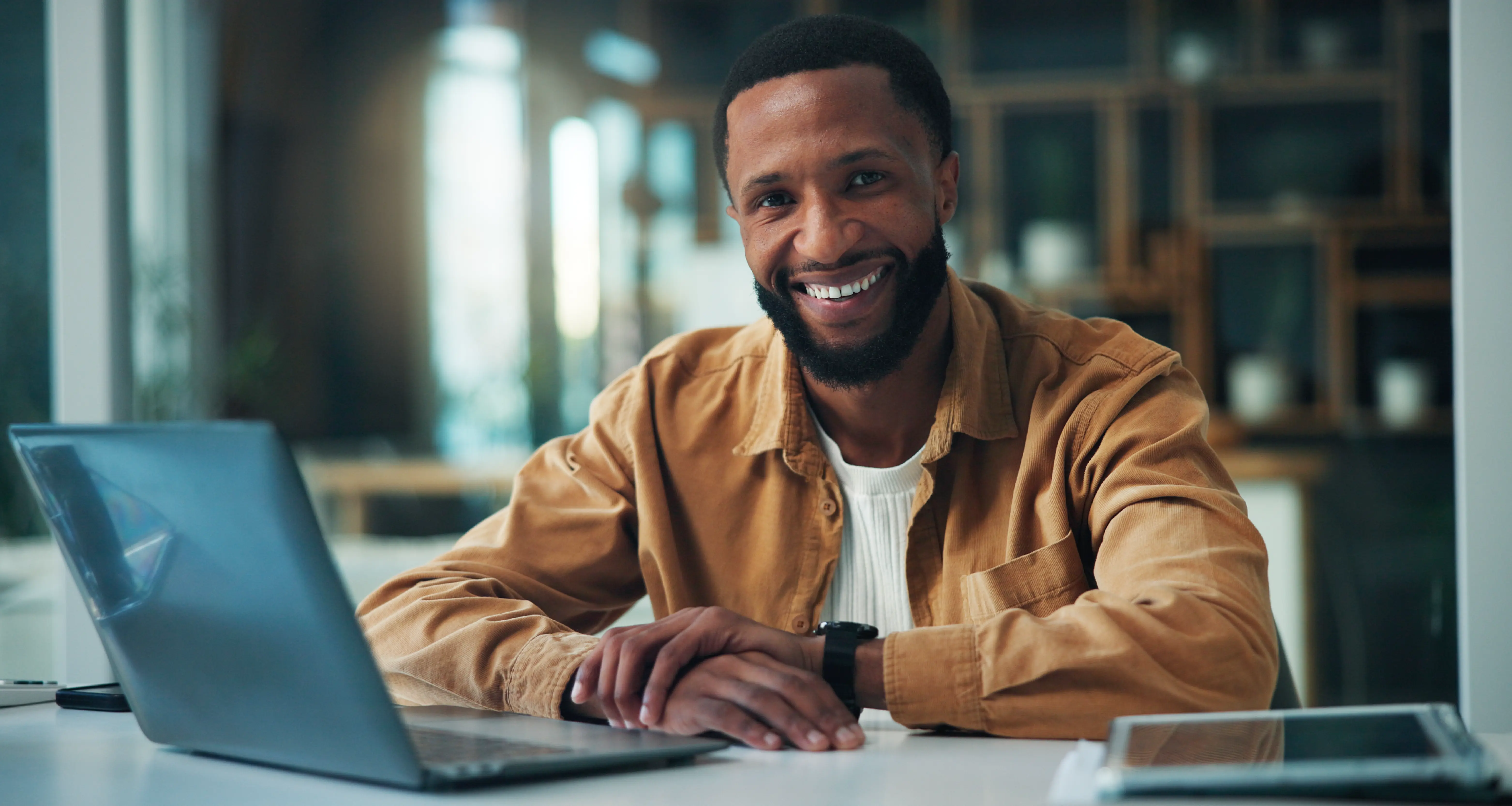 A smiling man with a beard wearing a tan jacket sits at a bright office desk with an open laptop, looking confidently at the camera, representing a skilled remote professional from South Africa available for hire by US companies.