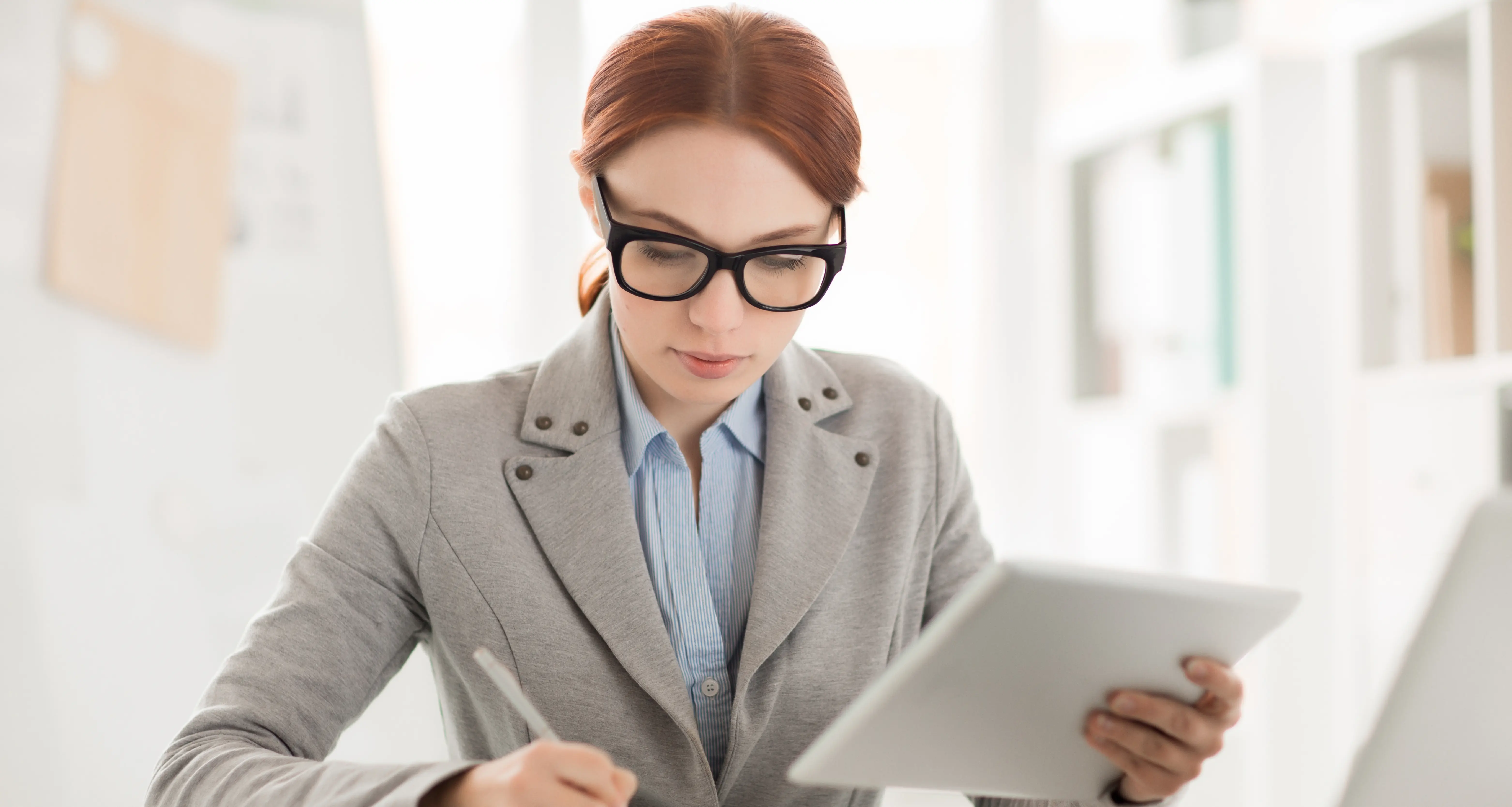 Red-haired woman in gray blazer and glasses intently reviewing content on a tablet while holding a pen in a bright office