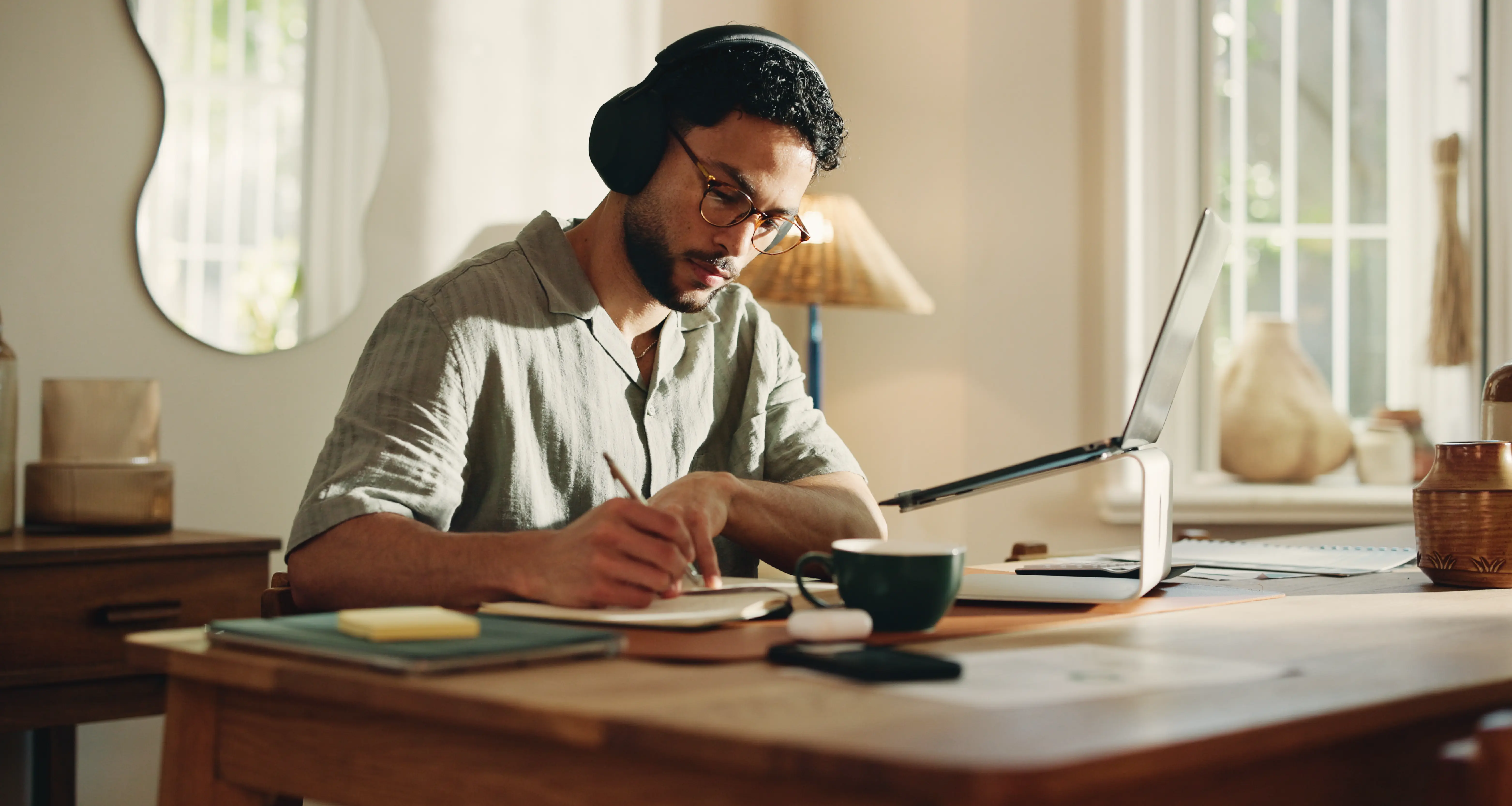 Man wearing headphones writing notes at a wooden desk with a laptop and coffee cup in a bright home workspace