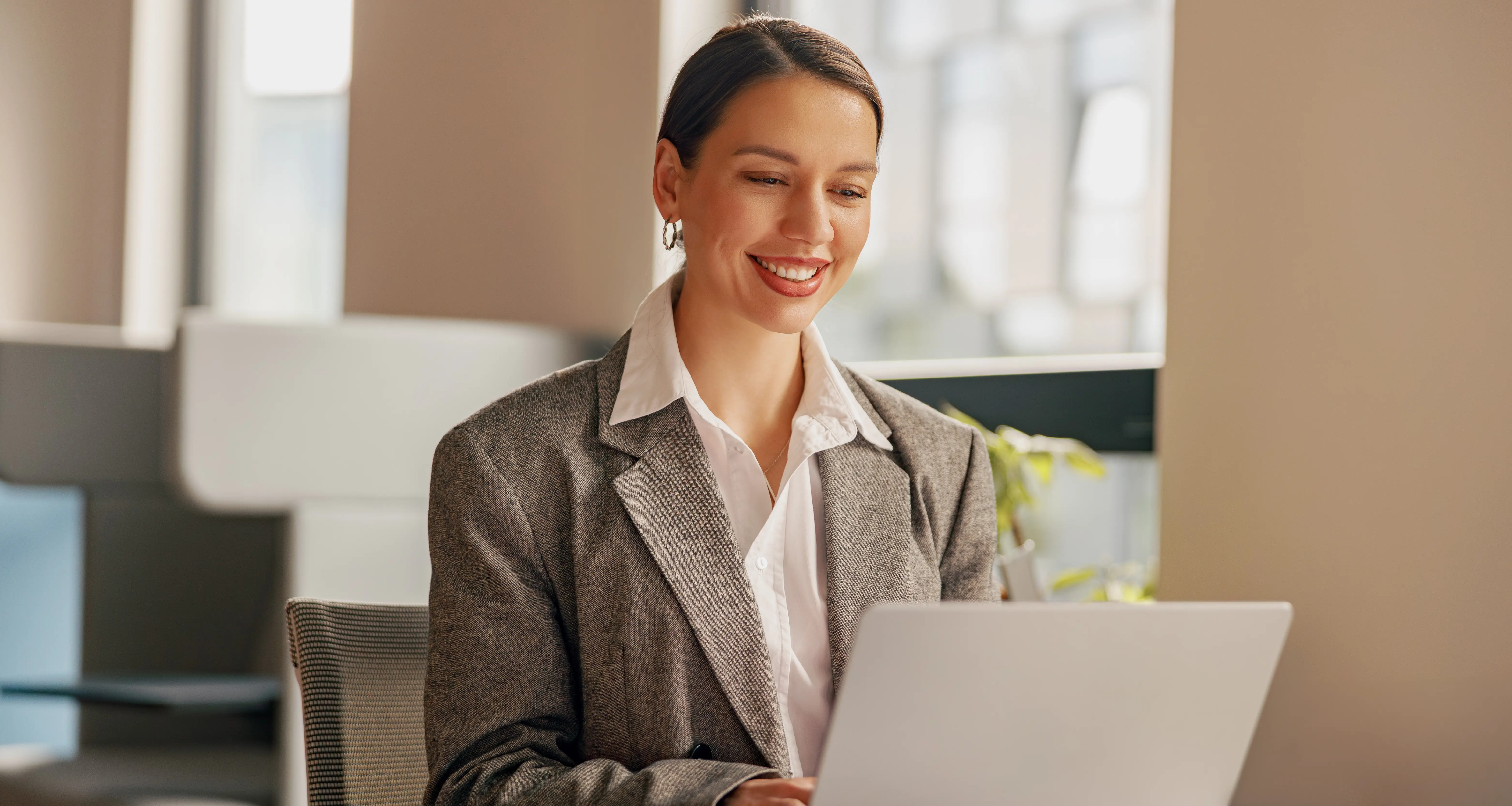 Smiling businesswoman in a gray blazer working on a laptop in a modern office environment
