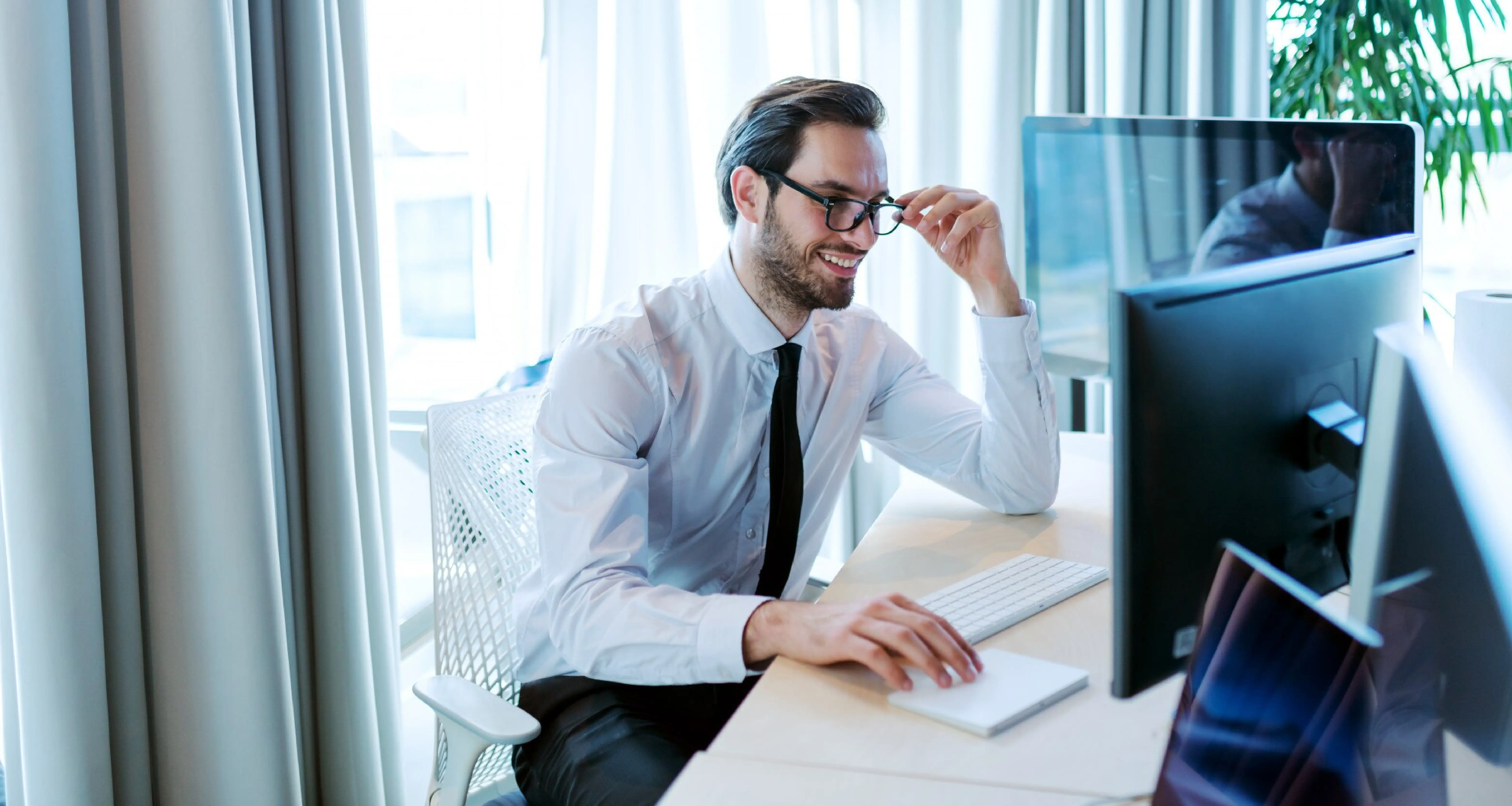 Businessman in a white shirt and tie adjusting his glasses while reviewing work on a desktop monitor