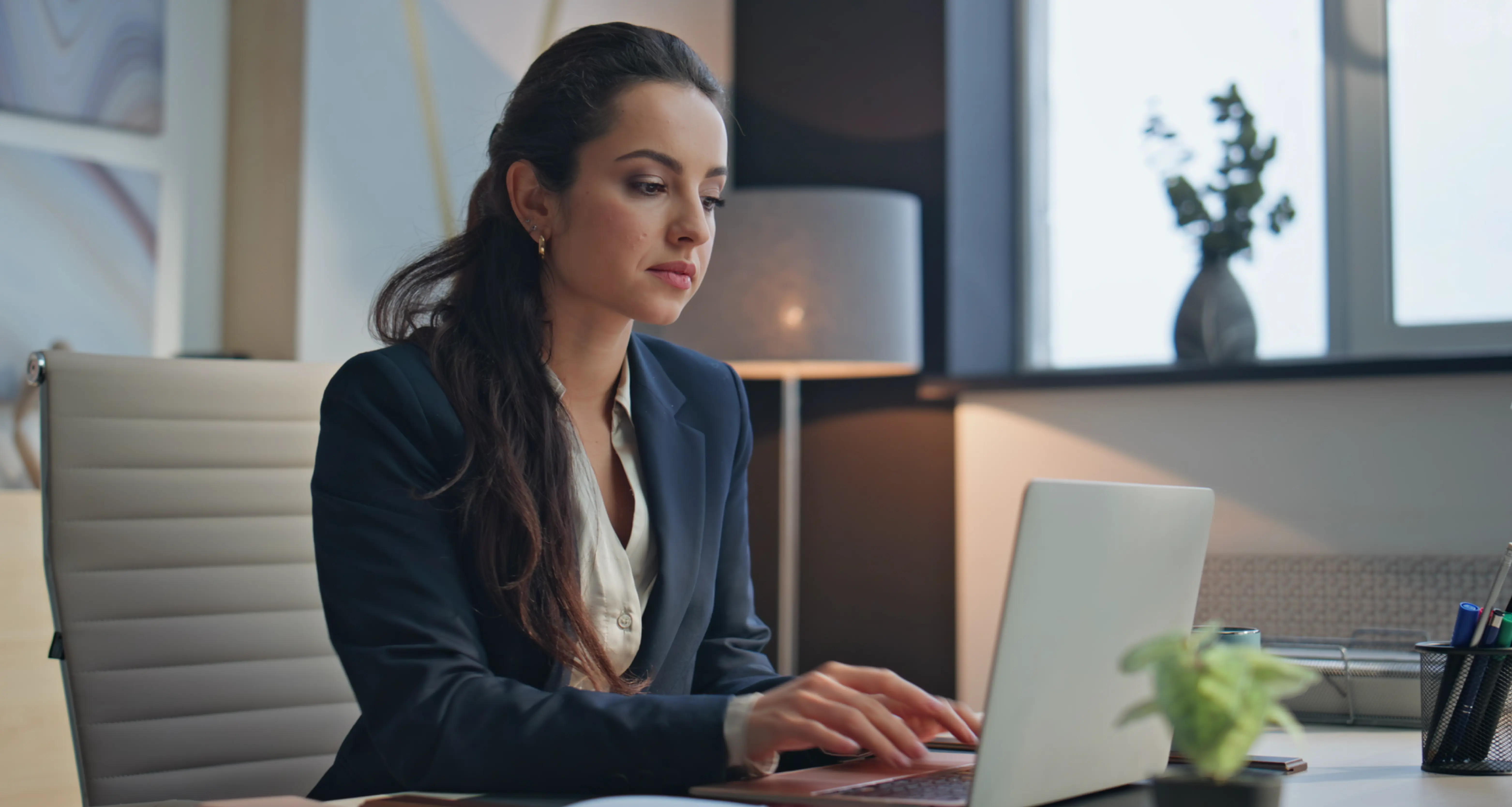 Professional woman in a dark blazer working on a laptop in a dimly lit modern office setting