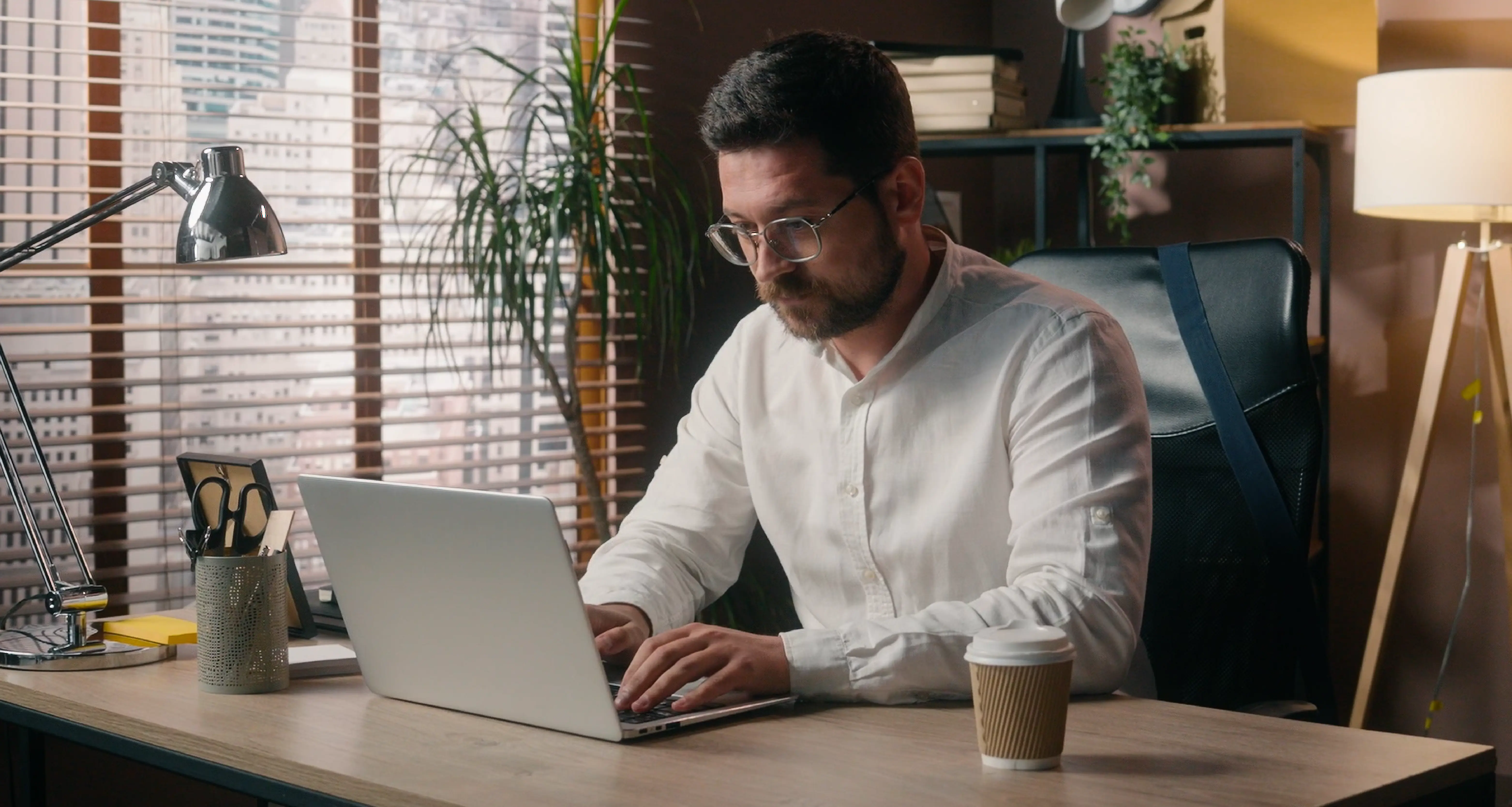 Man in glasses and white shirt typing on a laptop at a desk in a stylish office with city view through window blinds