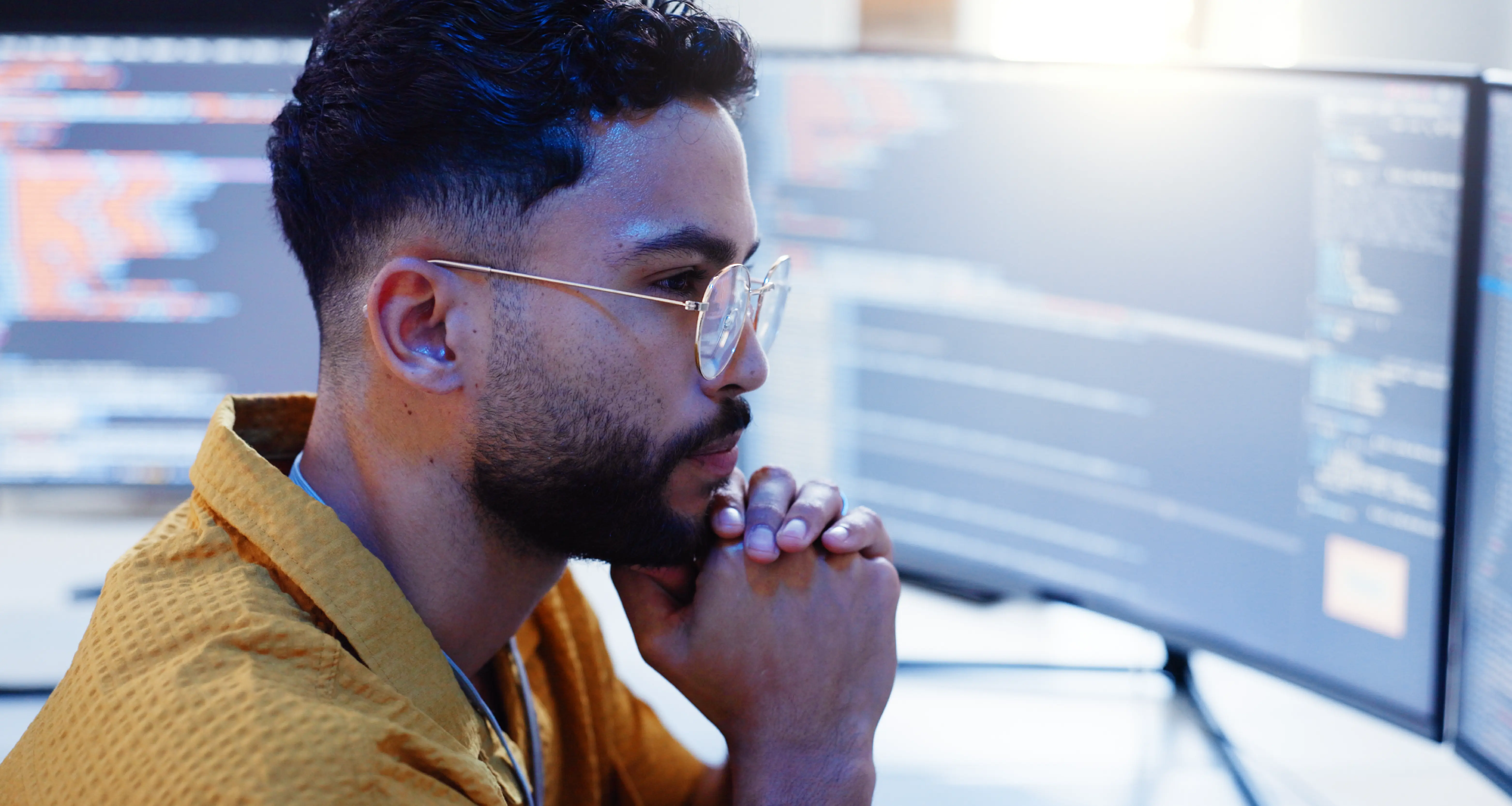 Developer with glasses and earbuds in contemplative focus in front of multiple monitors displaying code and data