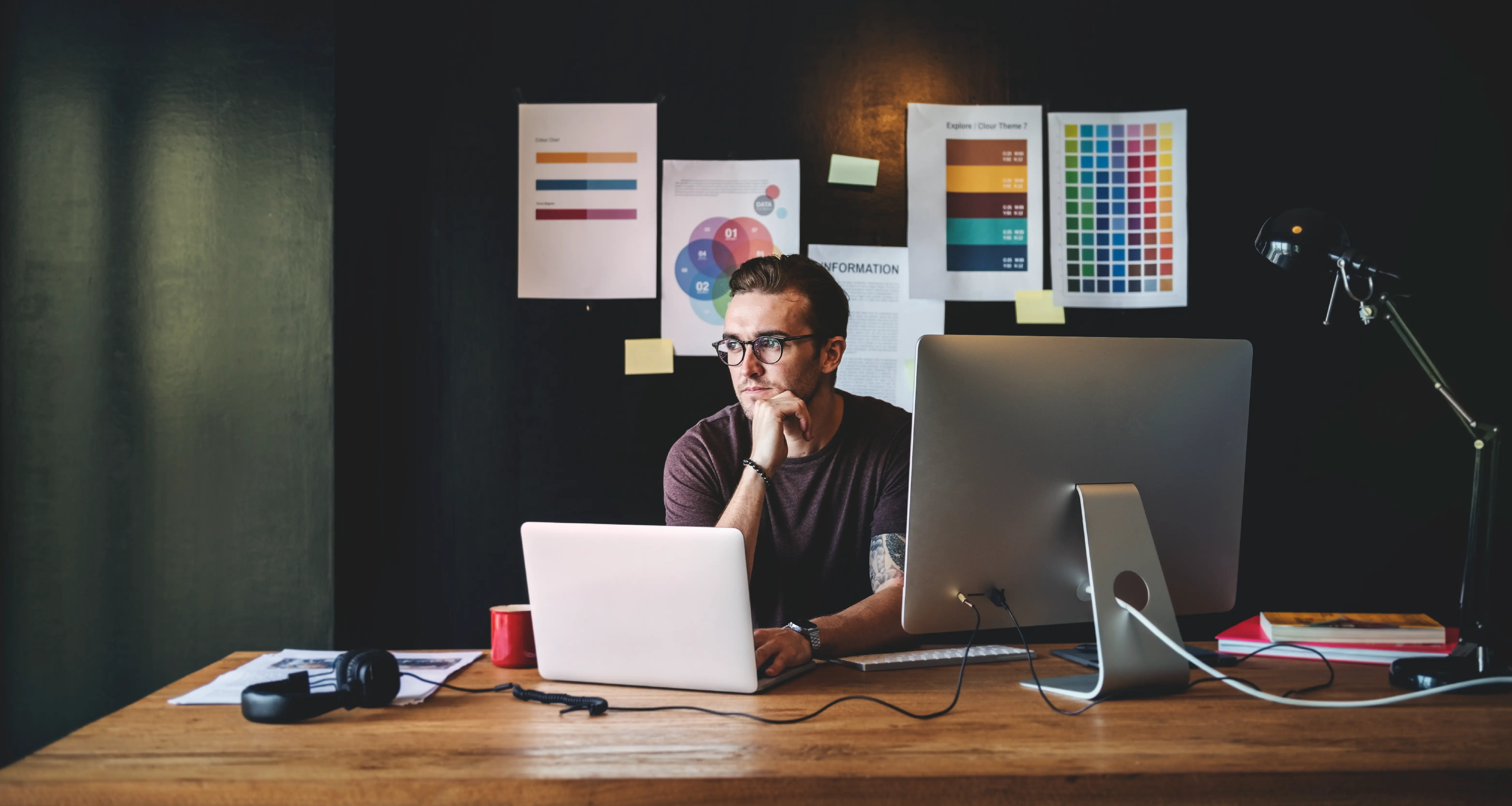 Creative designer with glasses working on dual monitors surrounded by color palettes and design references pinned to a dark wall