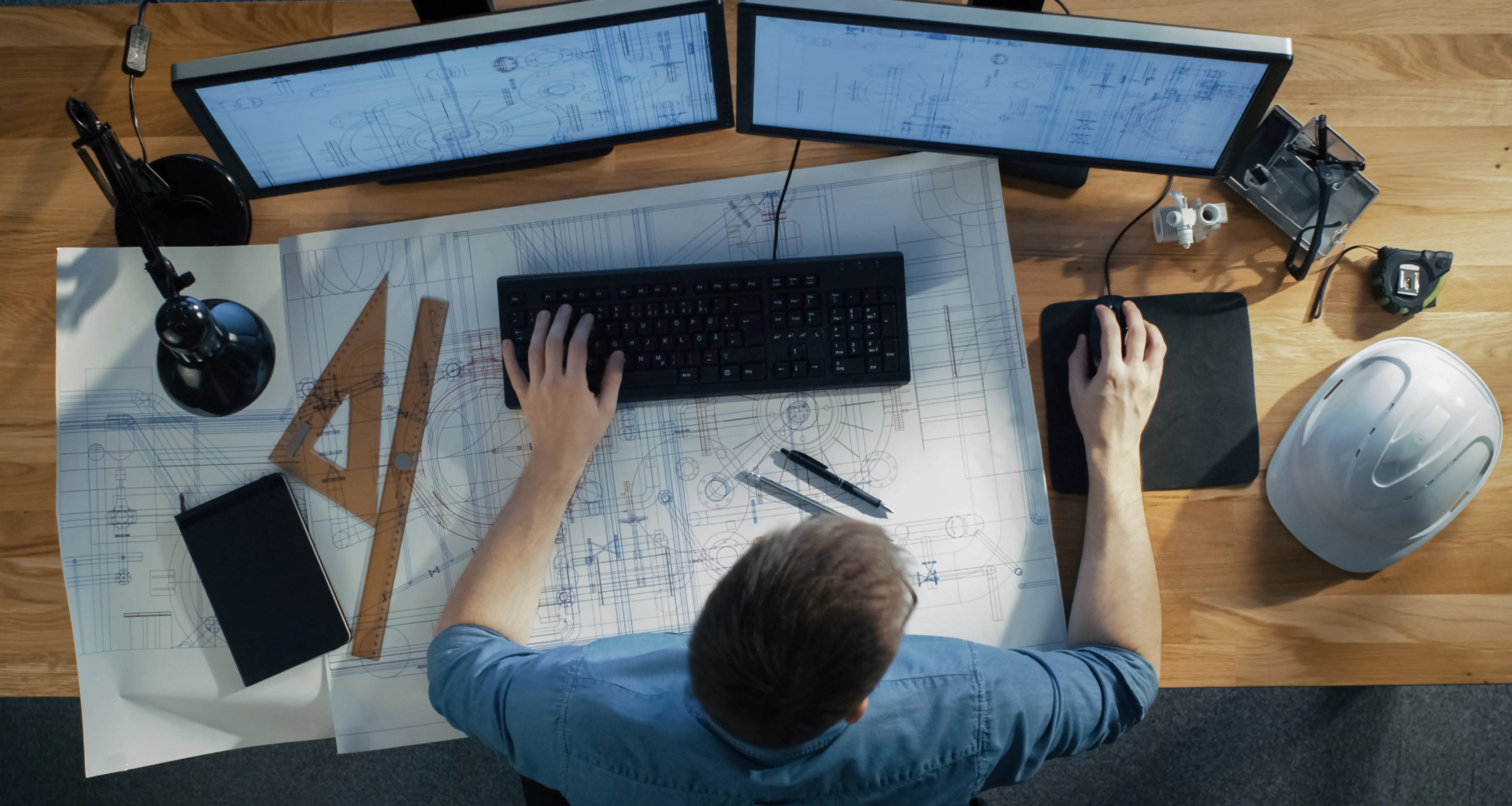 Overhead view of an engineer working on large architectural blueprints spread across a desk with dual monitors and a hard hat