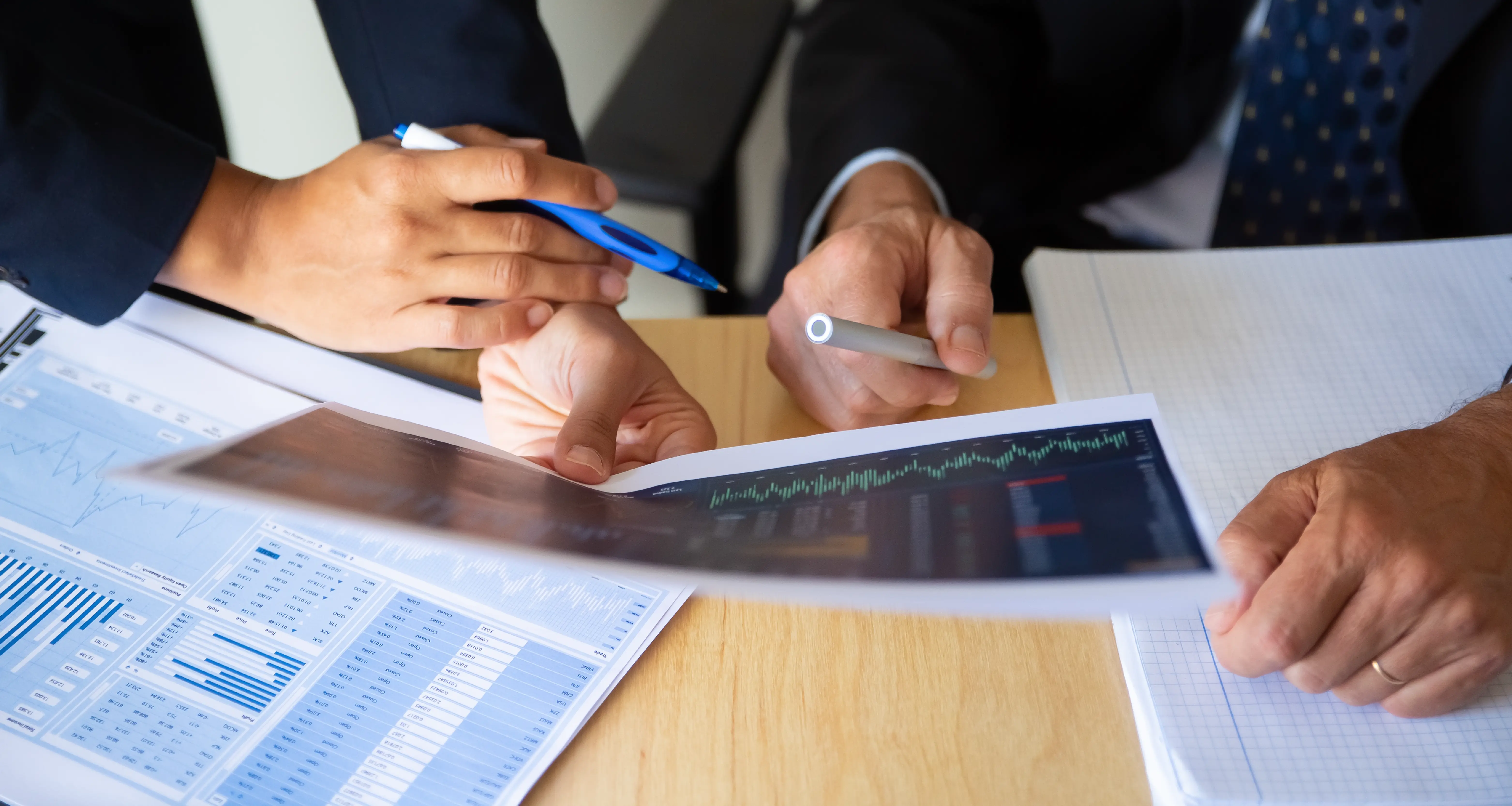 Two suited professionals reviewing financial charts and stock data on printed reports and a tablet at a wooden desk