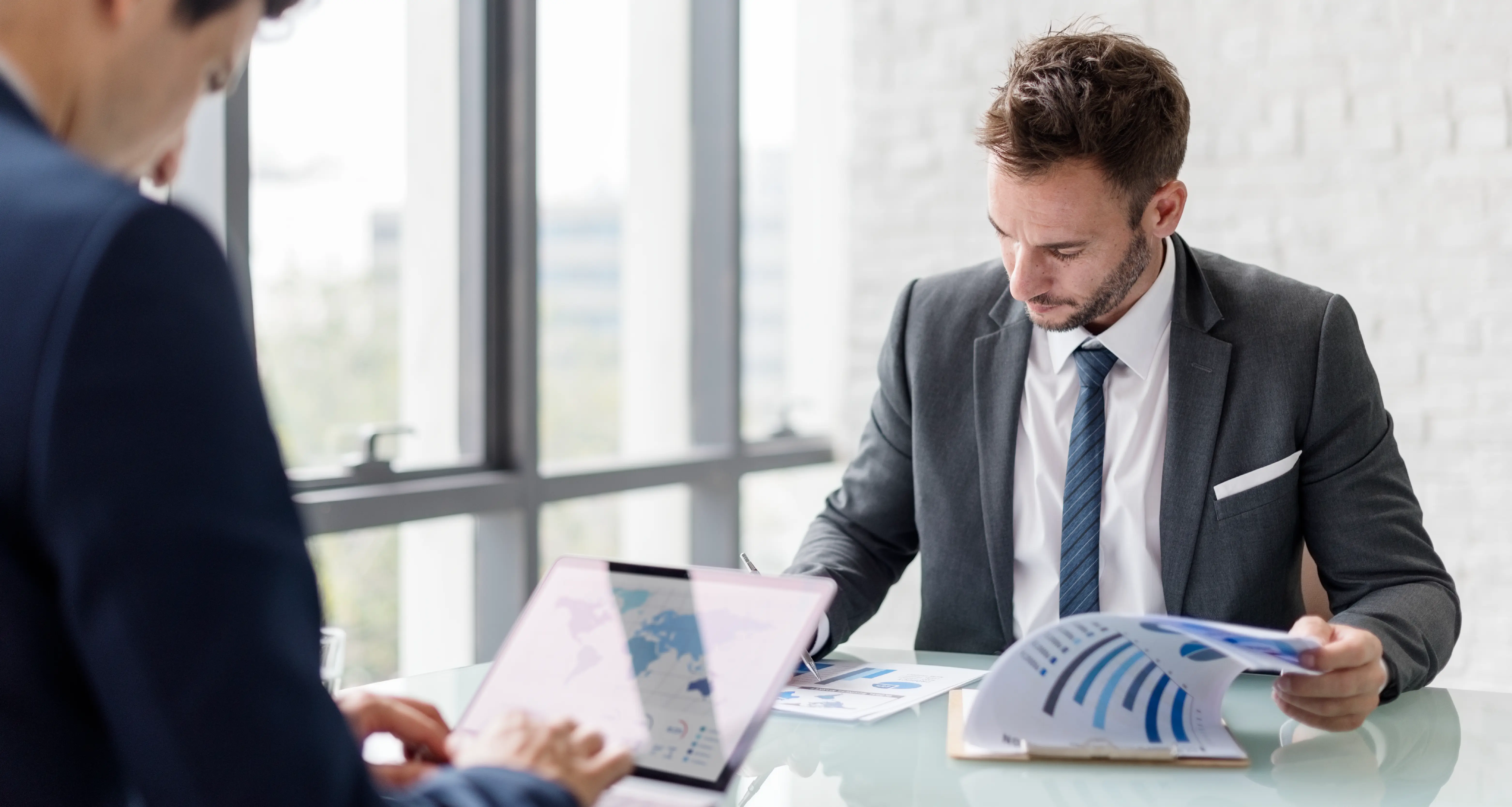 Businessman in gray suit reviewing a printed financial report at a glass table during a business meeting in a bright office