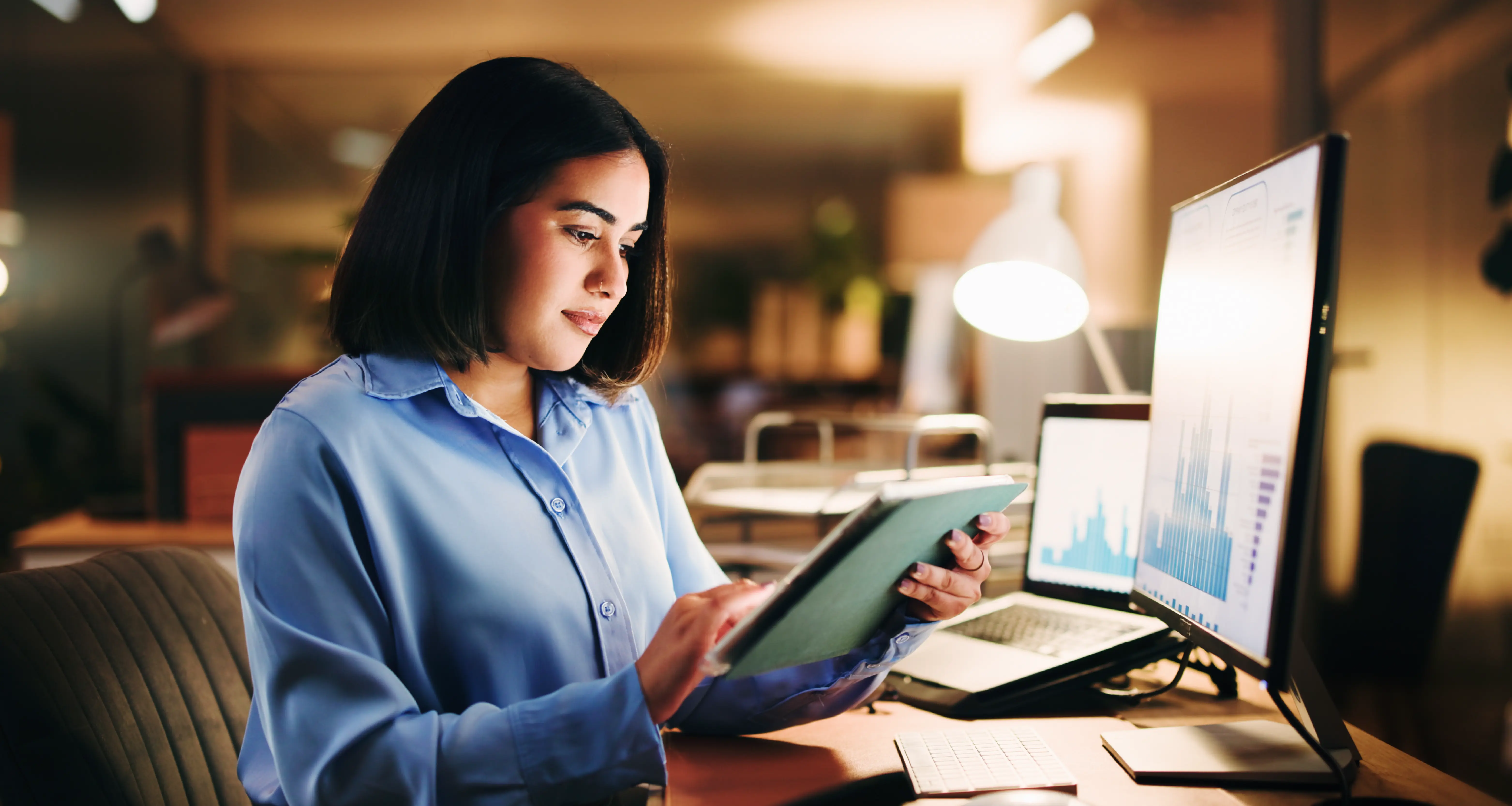 Woman in a blue shirt analyzing charts on a monitor and tablet at a desk in a dimly lit office at night