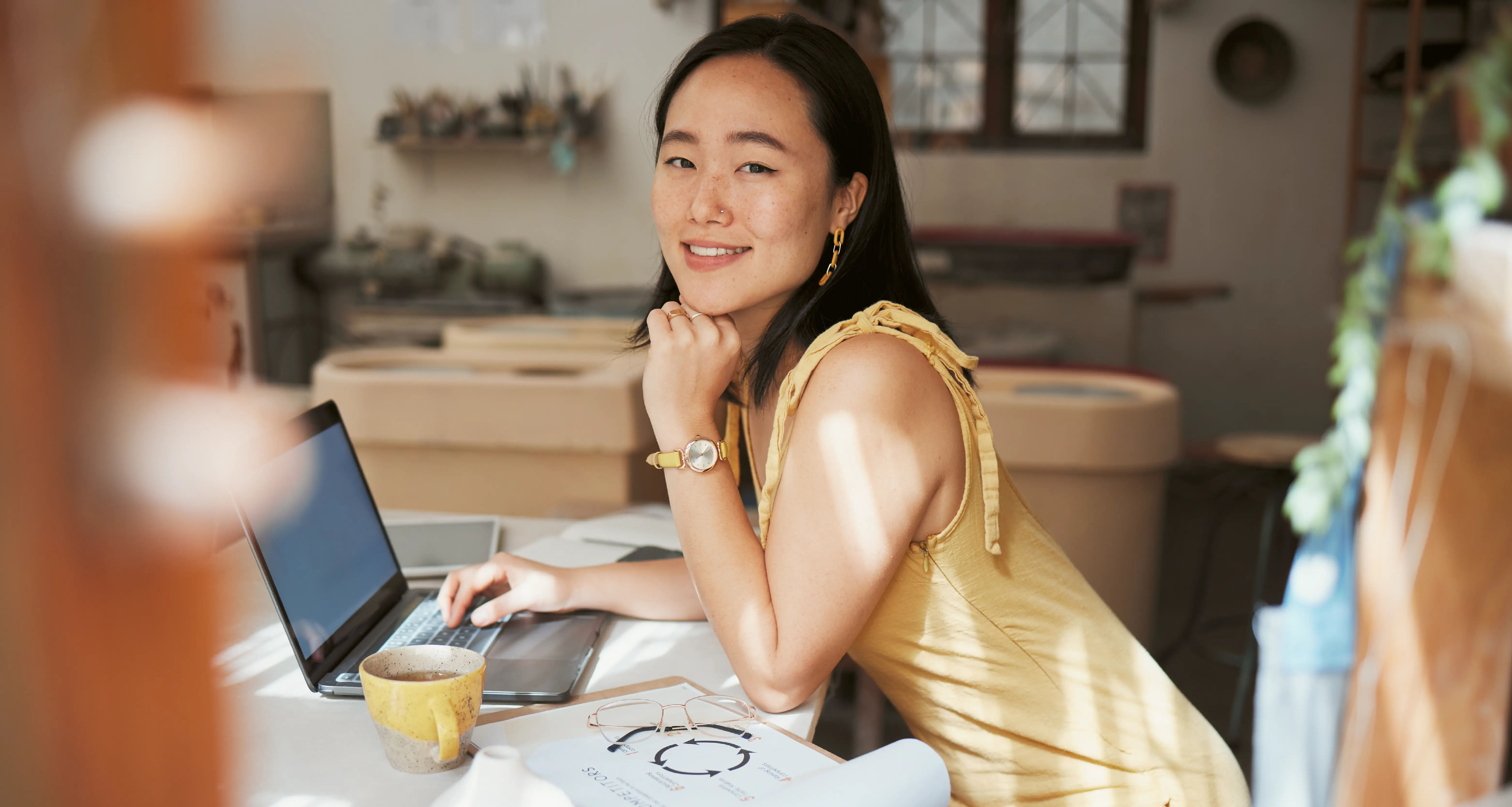 Young woman in a yellow dress working on a laptop with a coffee cup and notes in a cozy home workspace
