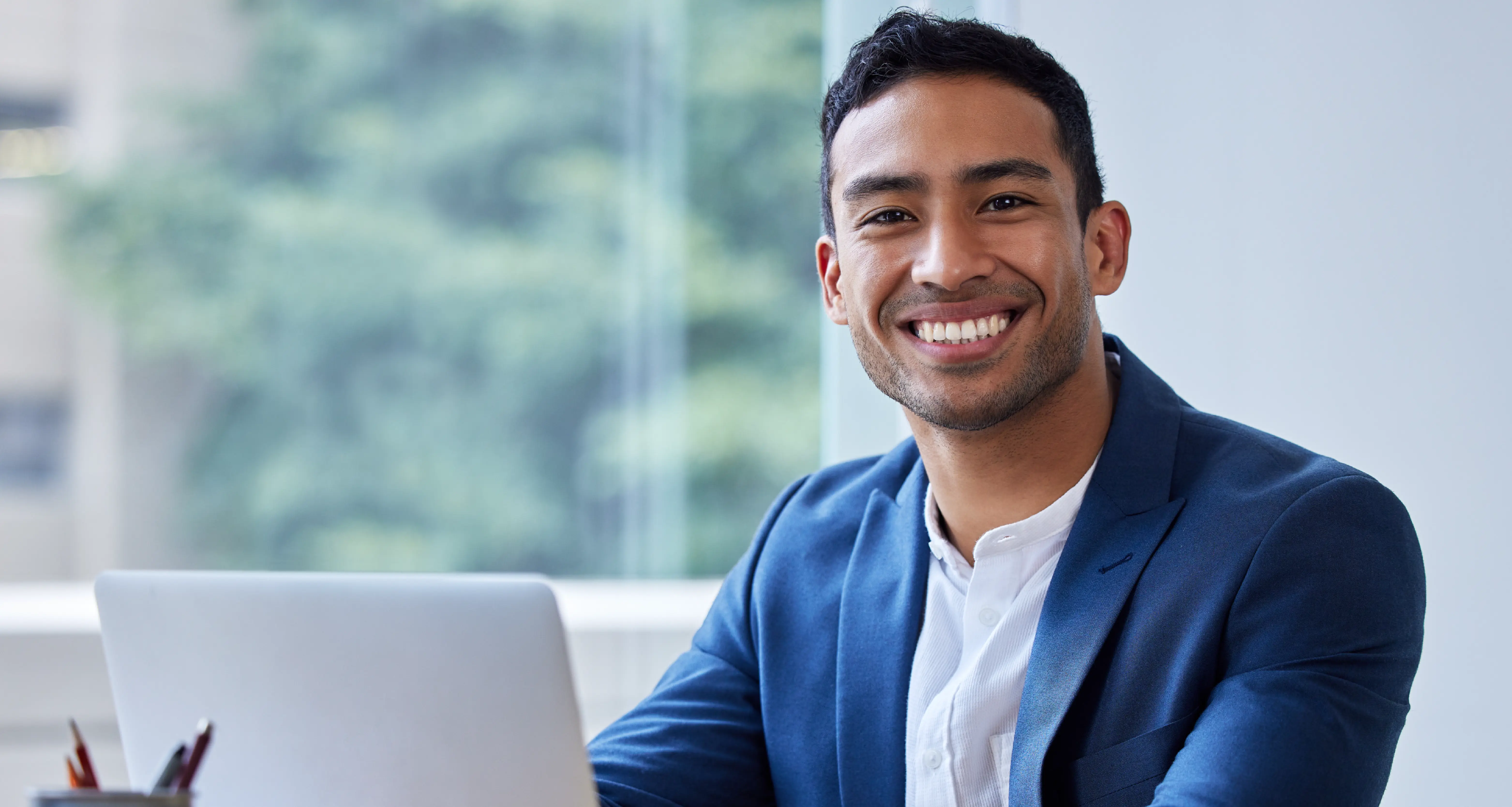 Confident young professional man in a navy blazer smiling at the camera while seated at a laptop in an office