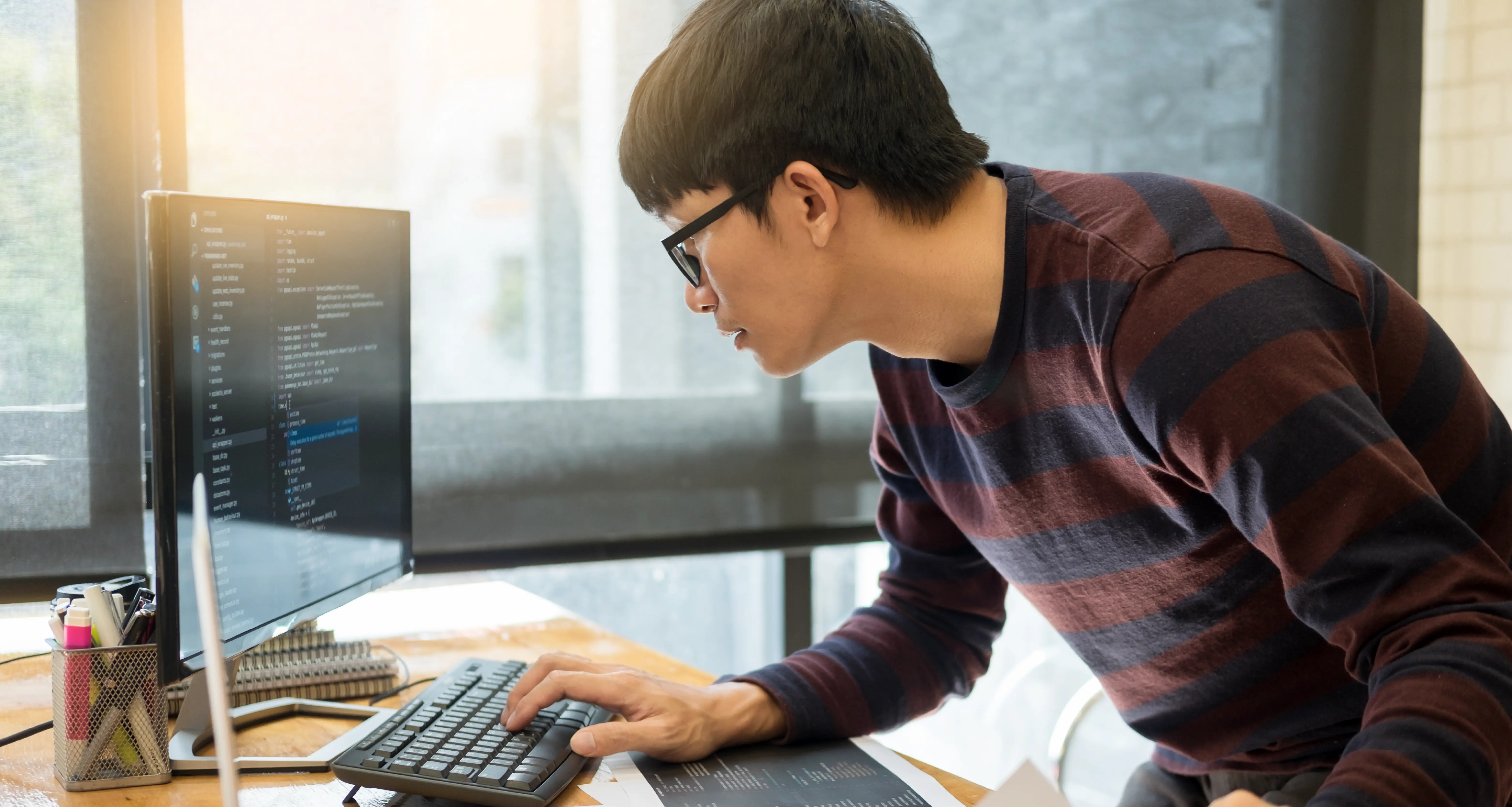 Developer with glasses leaning toward a monitor displaying code in a bright workspace with a striped sweater