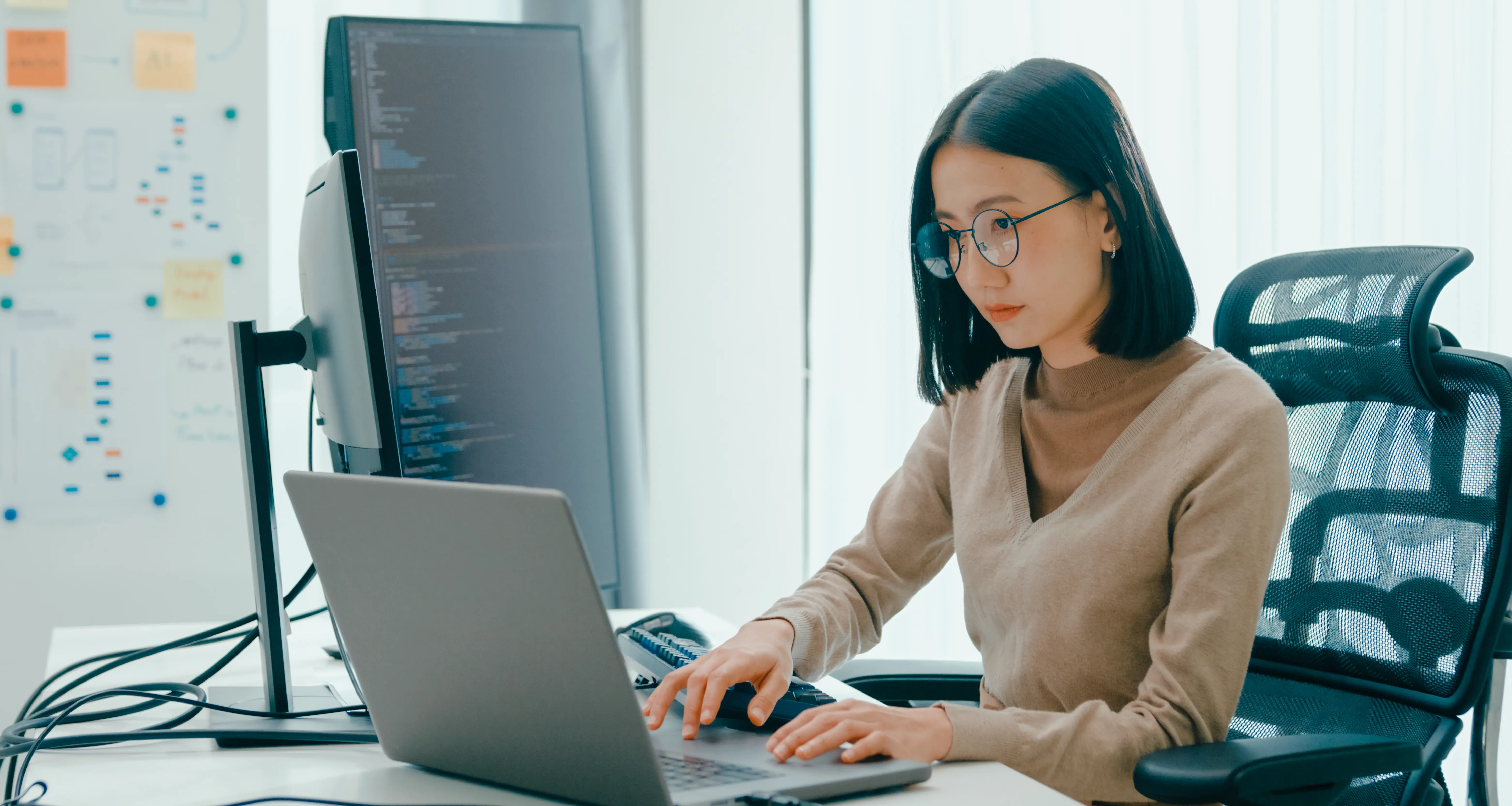 Female software engineer with glasses focused on laptop code in a modern office with a large monitor and sticky notes on a board