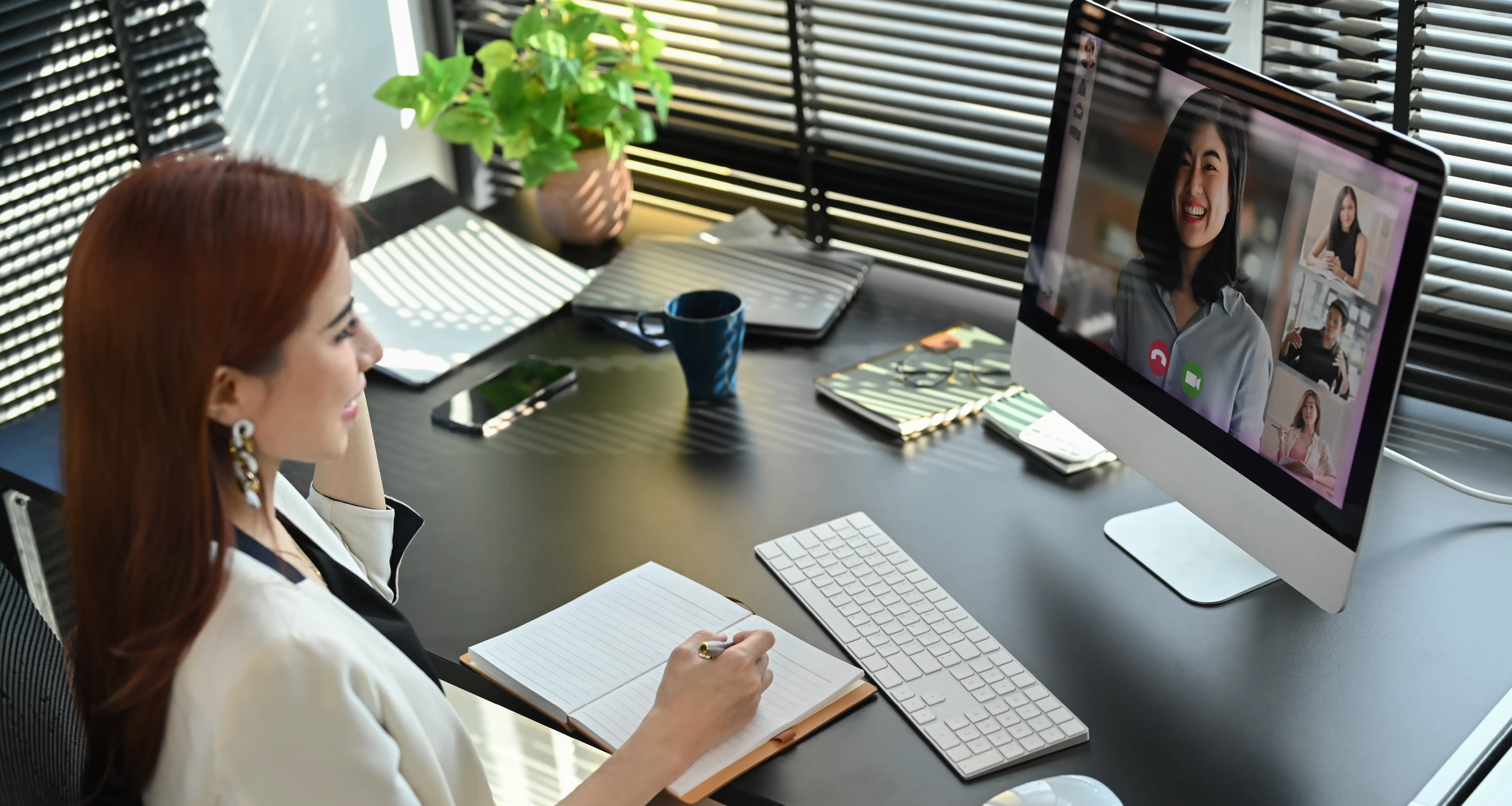 Remote worker attending a video call with multiple participants on a desktop screen while taking notes in a home office