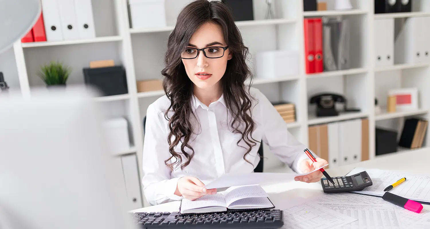 Professional woman with glasses reviewing documents and using a calculator at a tidy office workstation with binders on shelves