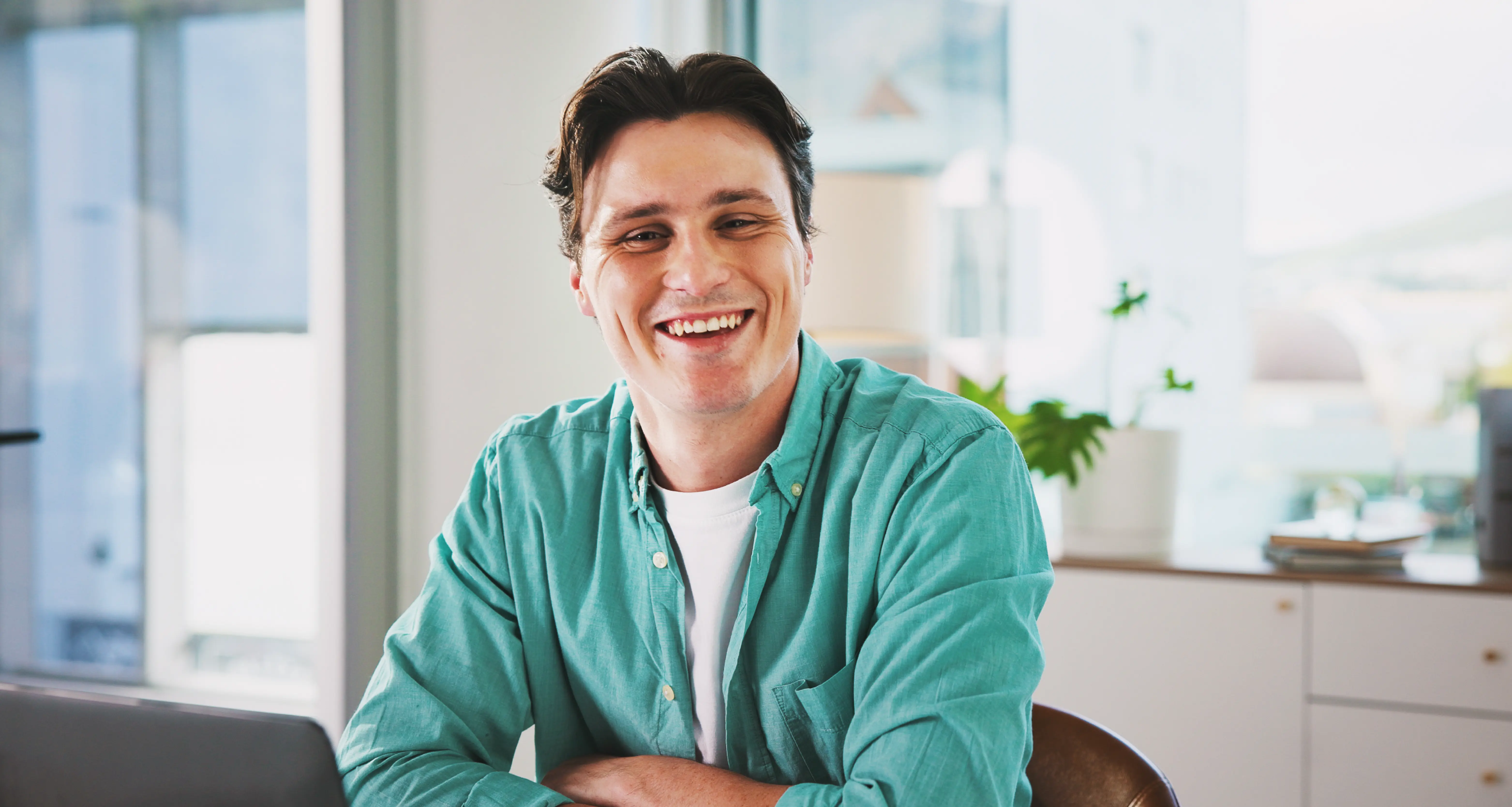 Smiling man in a teal shirt sitting at a desk with a laptop in a bright modern office
