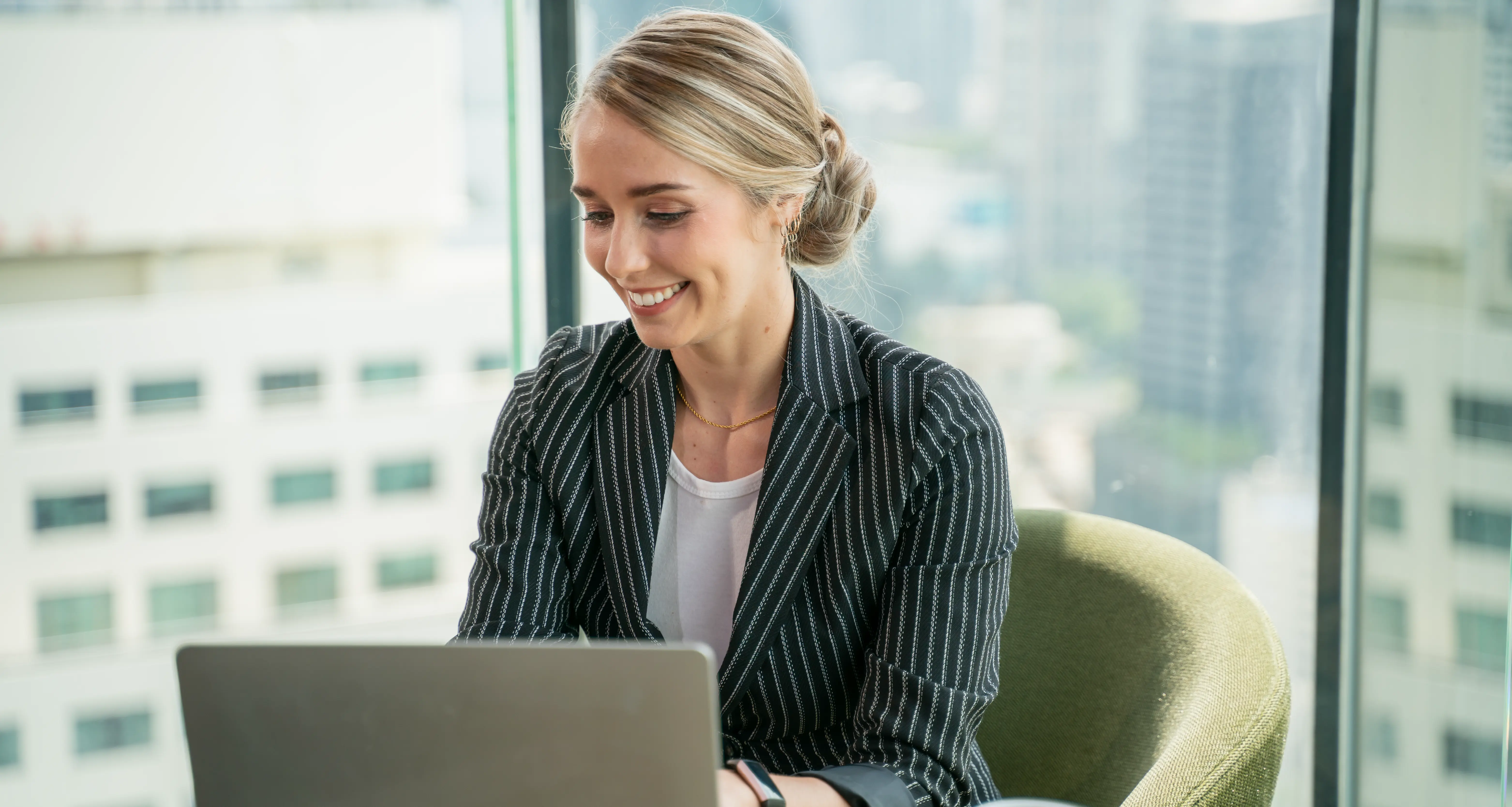 Blonde businesswoman using a laptop at a desk with a city skyline view through the window