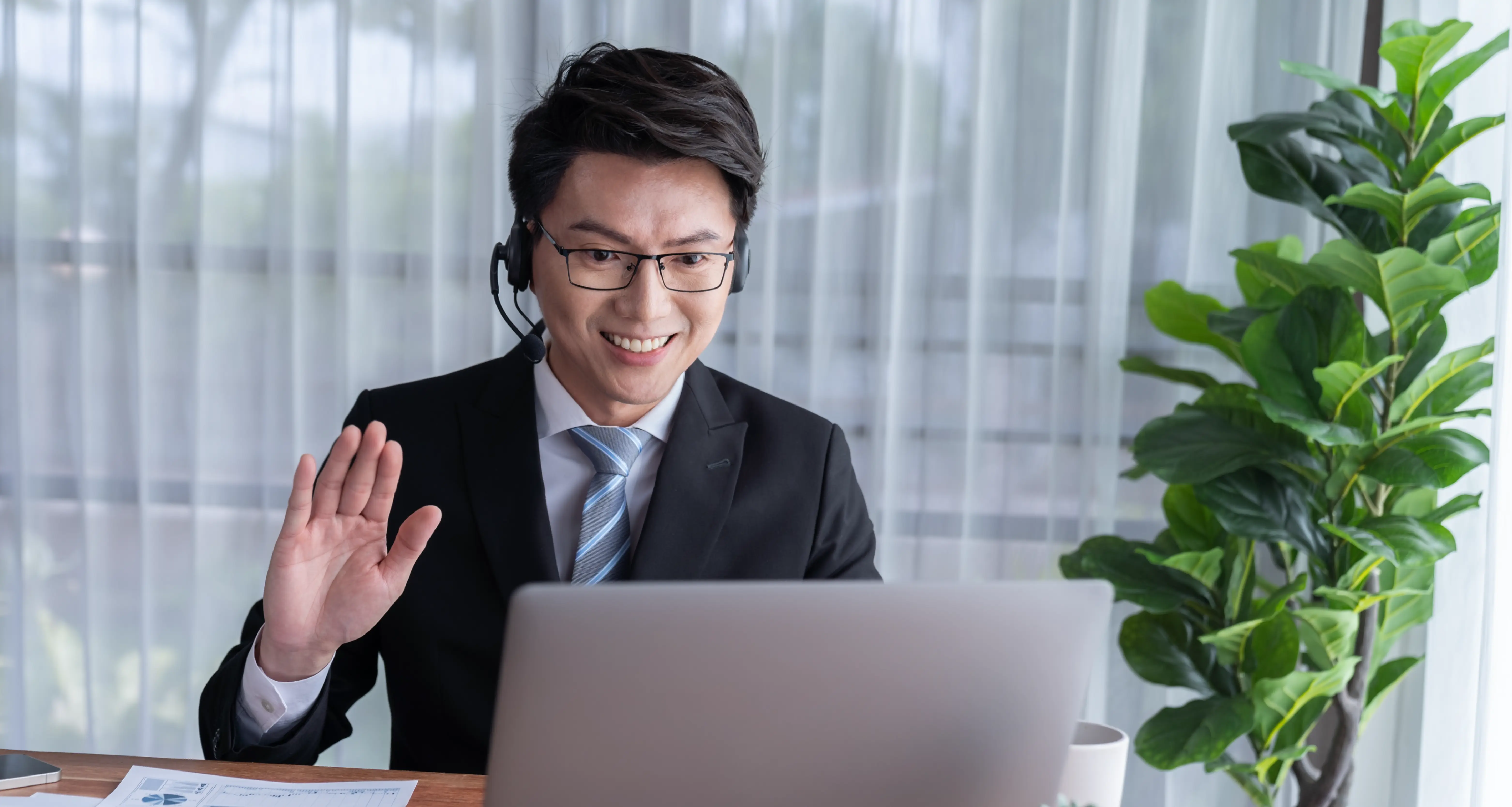 Businessman in a suit waving at a laptop screen while sitting at a desk in a professional office