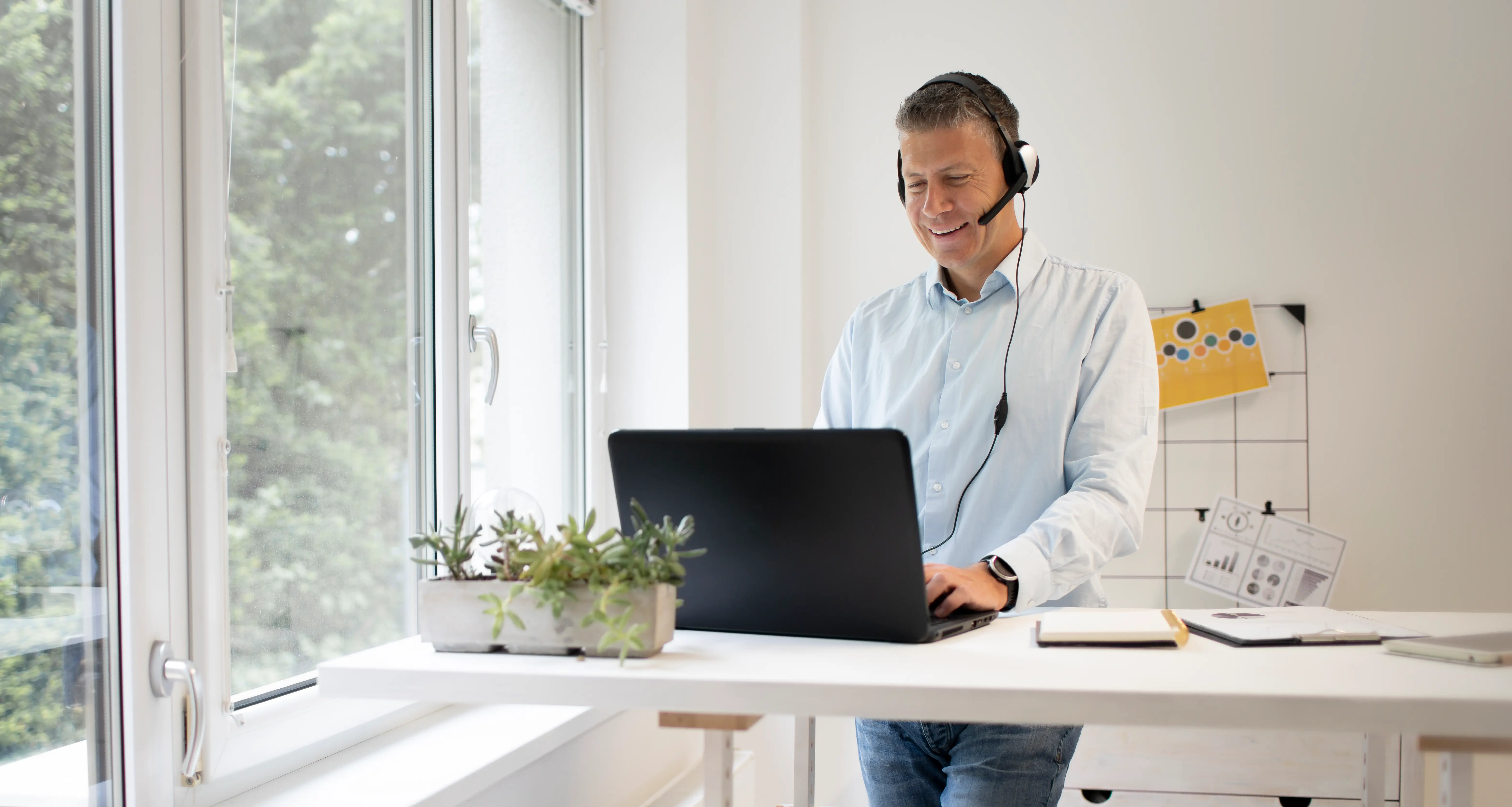Smiling man with a headset working on a laptop at a standing desk beside a bright window in a modern office