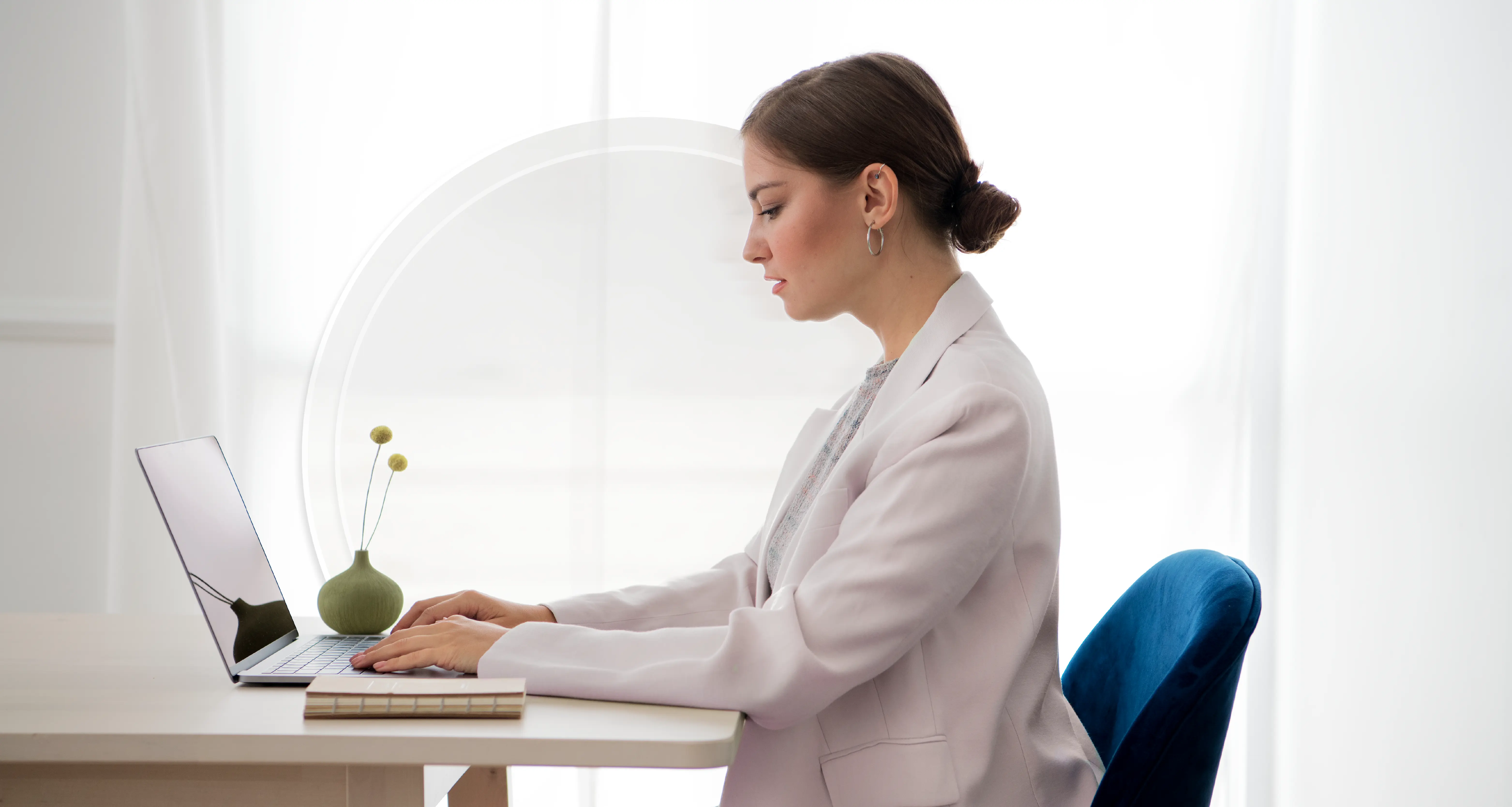 Professional woman in a white blazer focused on typing on a laptop at a minimalist white desk by a window