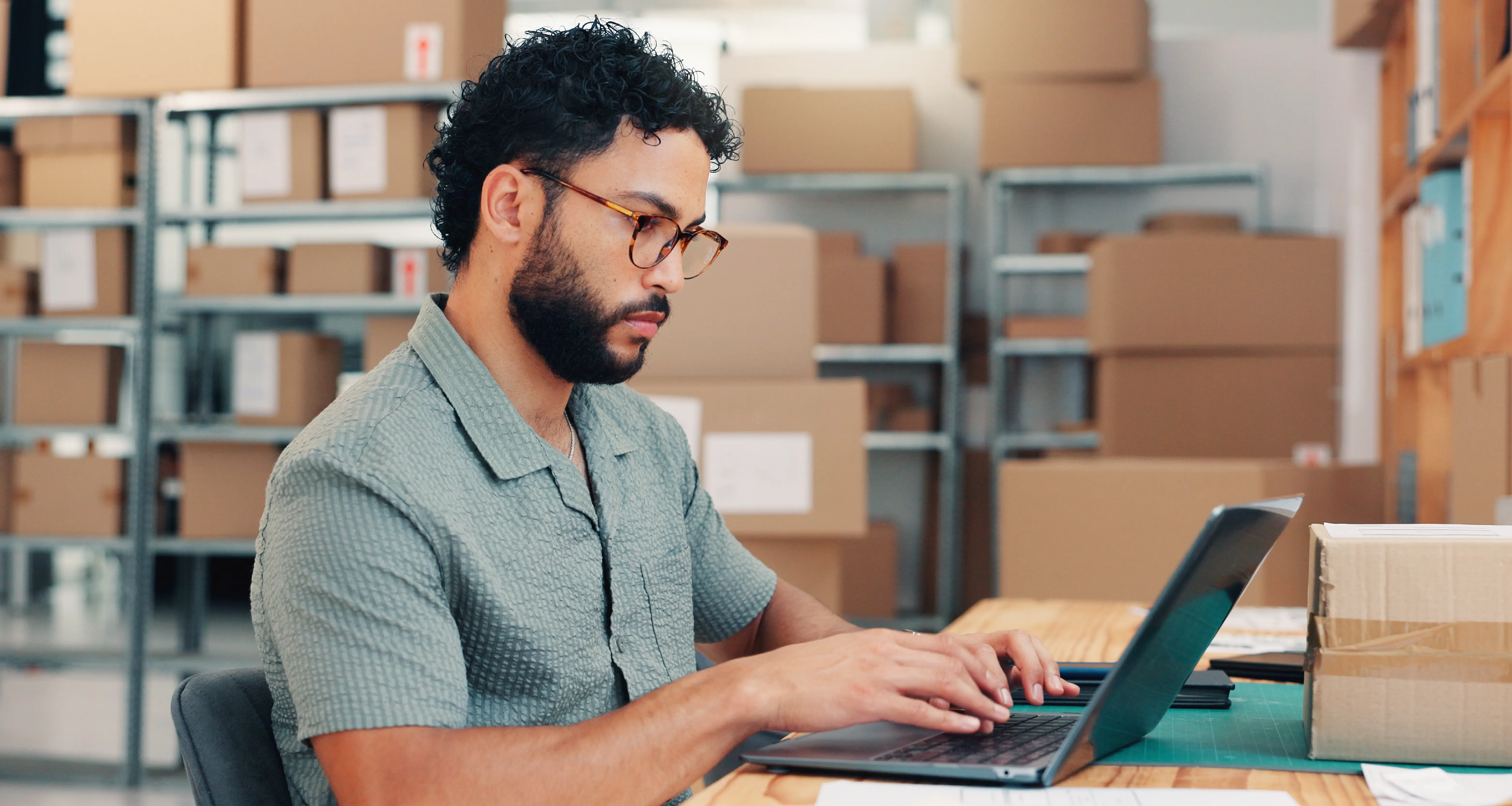 Man with curly hair and glasses working on a laptop in a warehouse surrounded by shelves of cardboard boxes