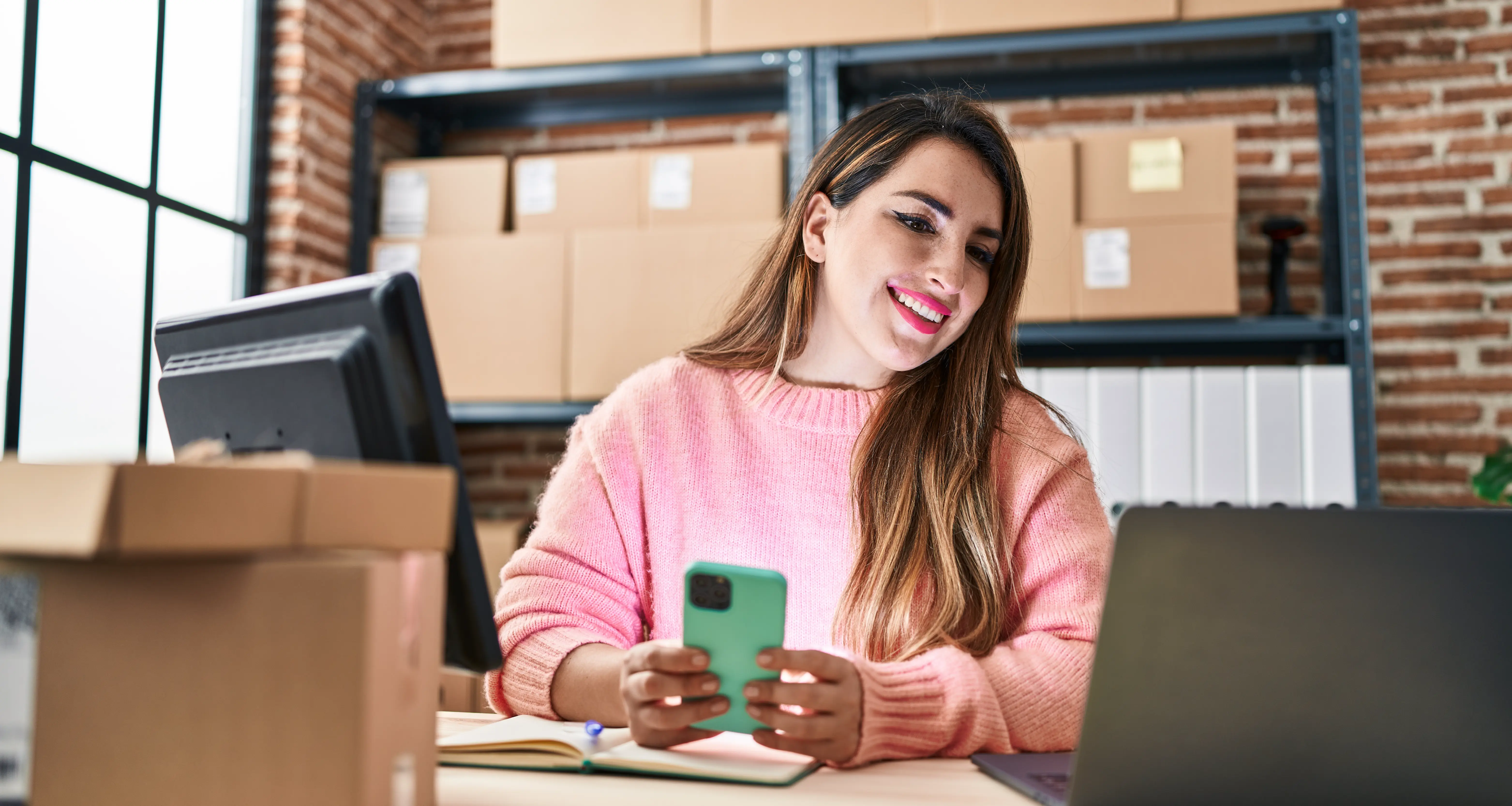 Smiling woman in a pink sweater holding a phone at a desk with a computer and shipping boxes in a warehouse office