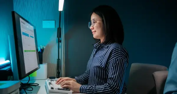 Woman with glasses working focused at a desktop computer in a dark blue-lit modern home office at night