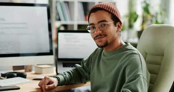 Young man with glasses and a beanie hat sitting at a dual-monitor desk with code on screen in a plant-filled home office