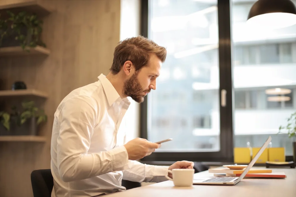 A caucasian man holding his phone whilst looking at his laptop. He's probably developing an email triage for his assistant.