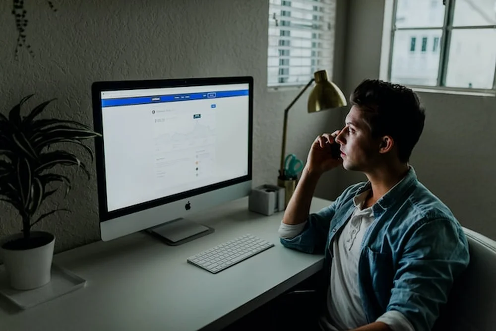 A man sitting in front of a computer talking on his cell phone